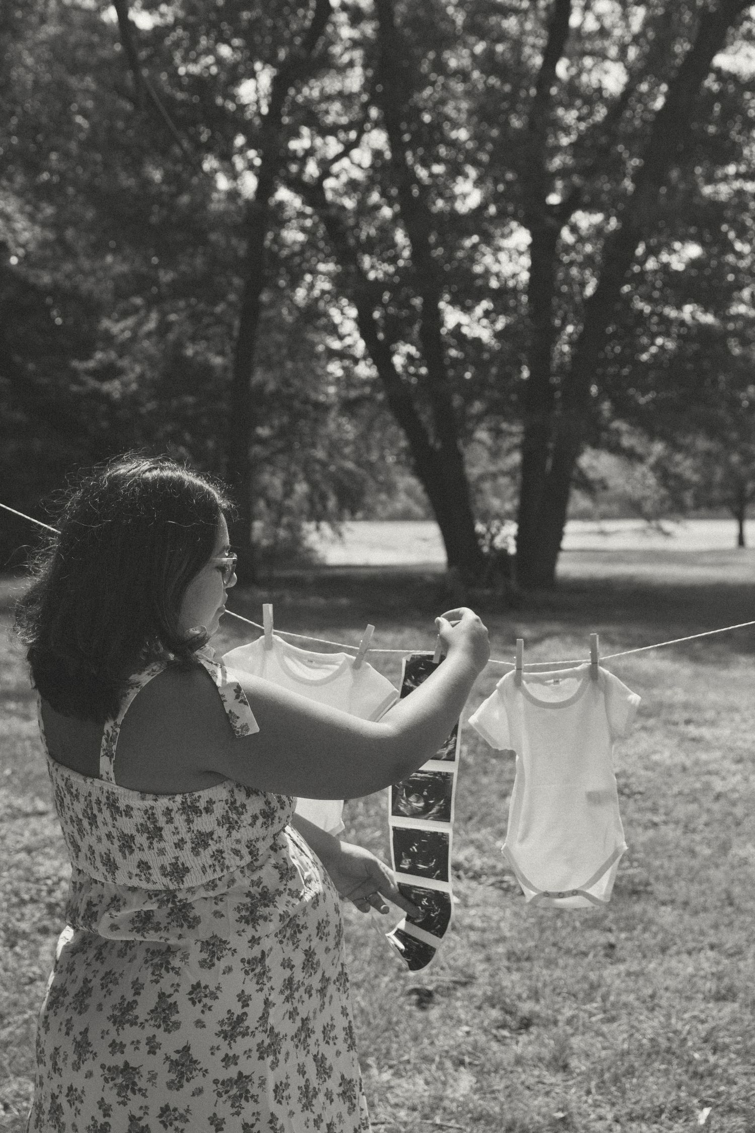 Pregnant woman in floral dress hangs baby onesies and ultrasound photos on a clothesline in a sunlit park, celebrating motherhood.