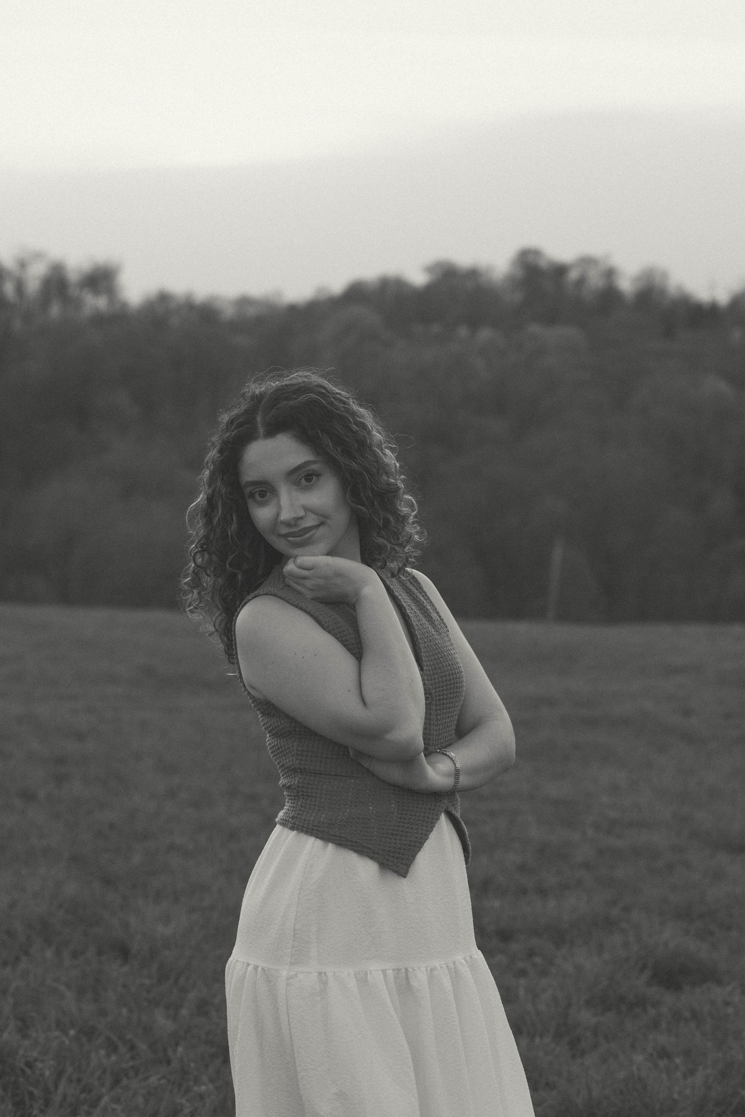 Debbie from D'Gallery Photography poses thoughtfully in a white, flowy skirt against a misty mountain backdrop in a grassy field.