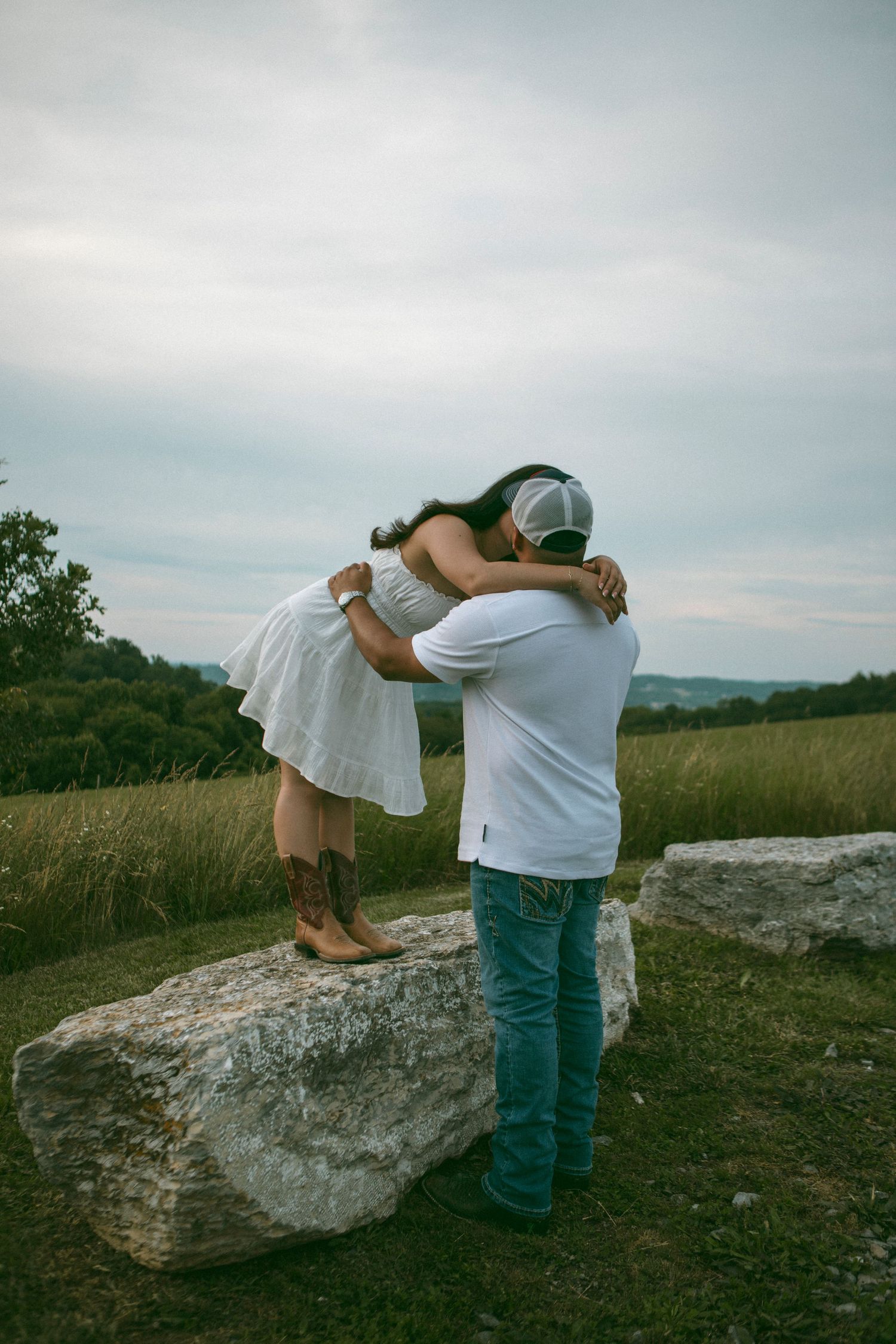 A couple shares a romantic moment on a large rock formation overlooking a scenic rural landscape at dusk.