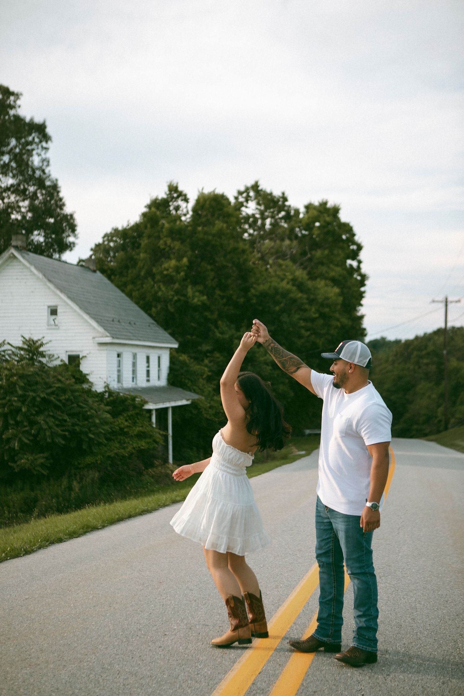A couple dances playfully together on an empty rural road with a white house visible in the distance during a storytelling couples session.