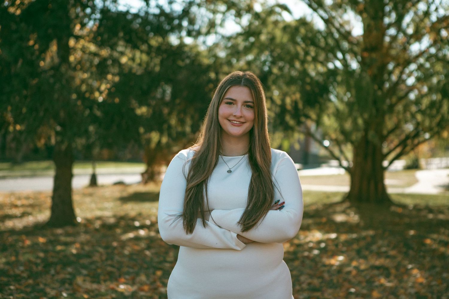 Confident young woman in a white dress posing with arms crossed in a park with autumn leaves and sunlight filtering through trees.