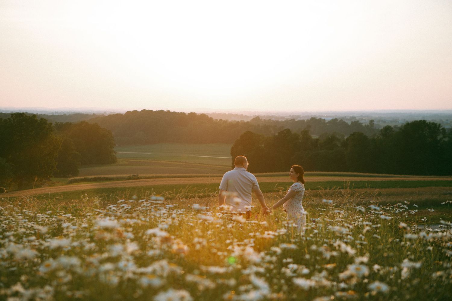 A couple walks through a sunlit meadow filled with white wildflowers during golden hour for a maternity storytelling session.