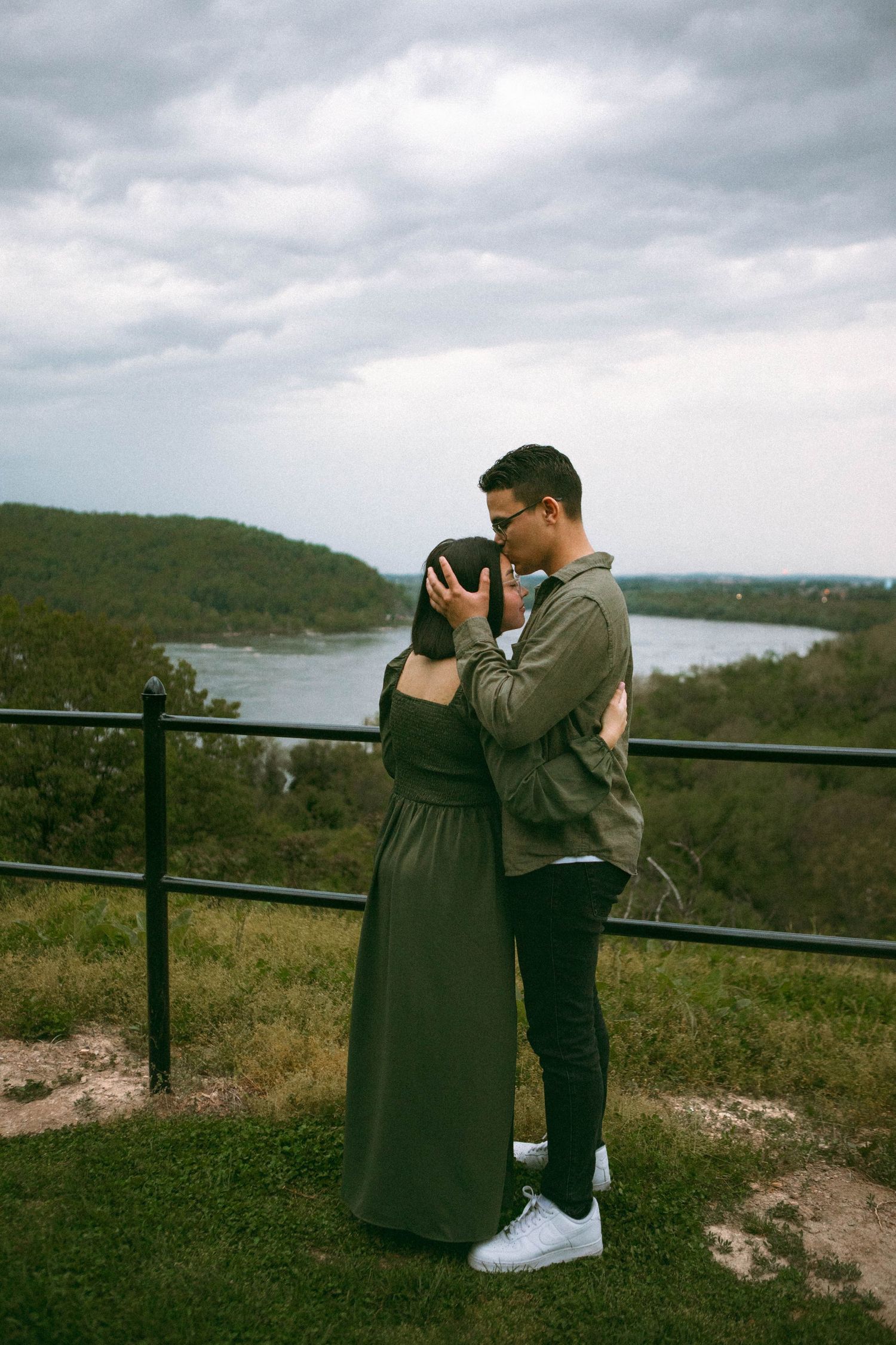 Couple embraces while the man gives the woman a forehead kiss, in front of a scenic river view after an engagement couples session.
