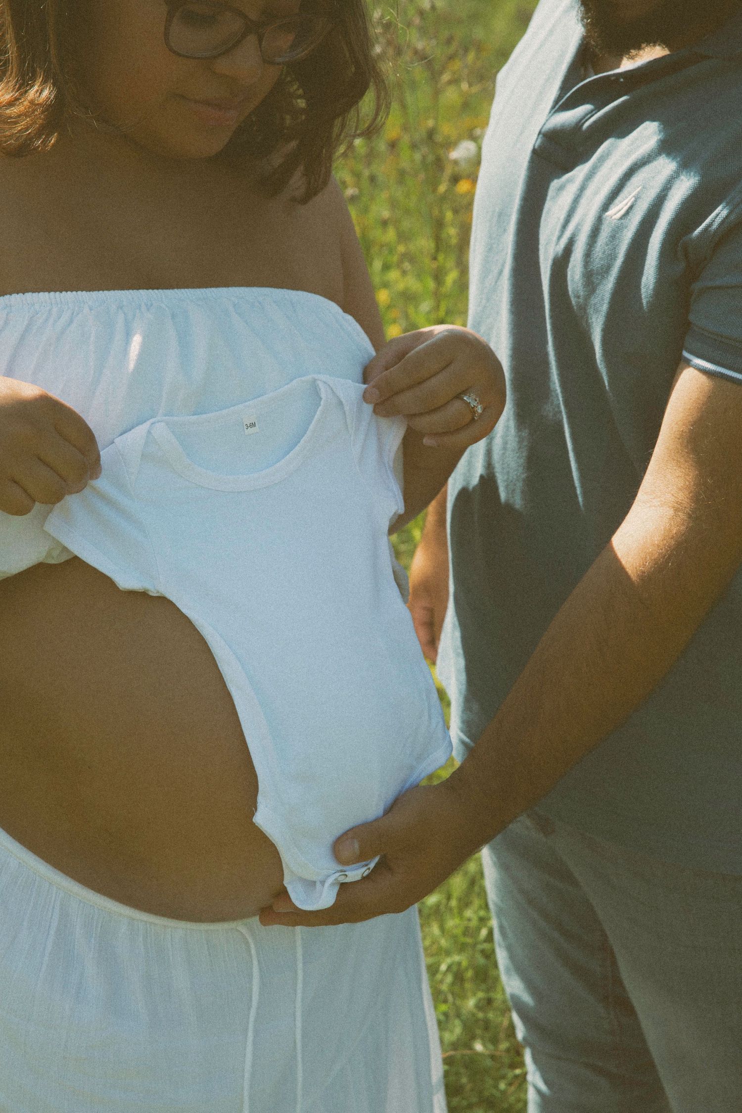Pregnant woman in a white dress holds a baby onesie over her belly while her partner supports her, capturing a joyful maternity announcement