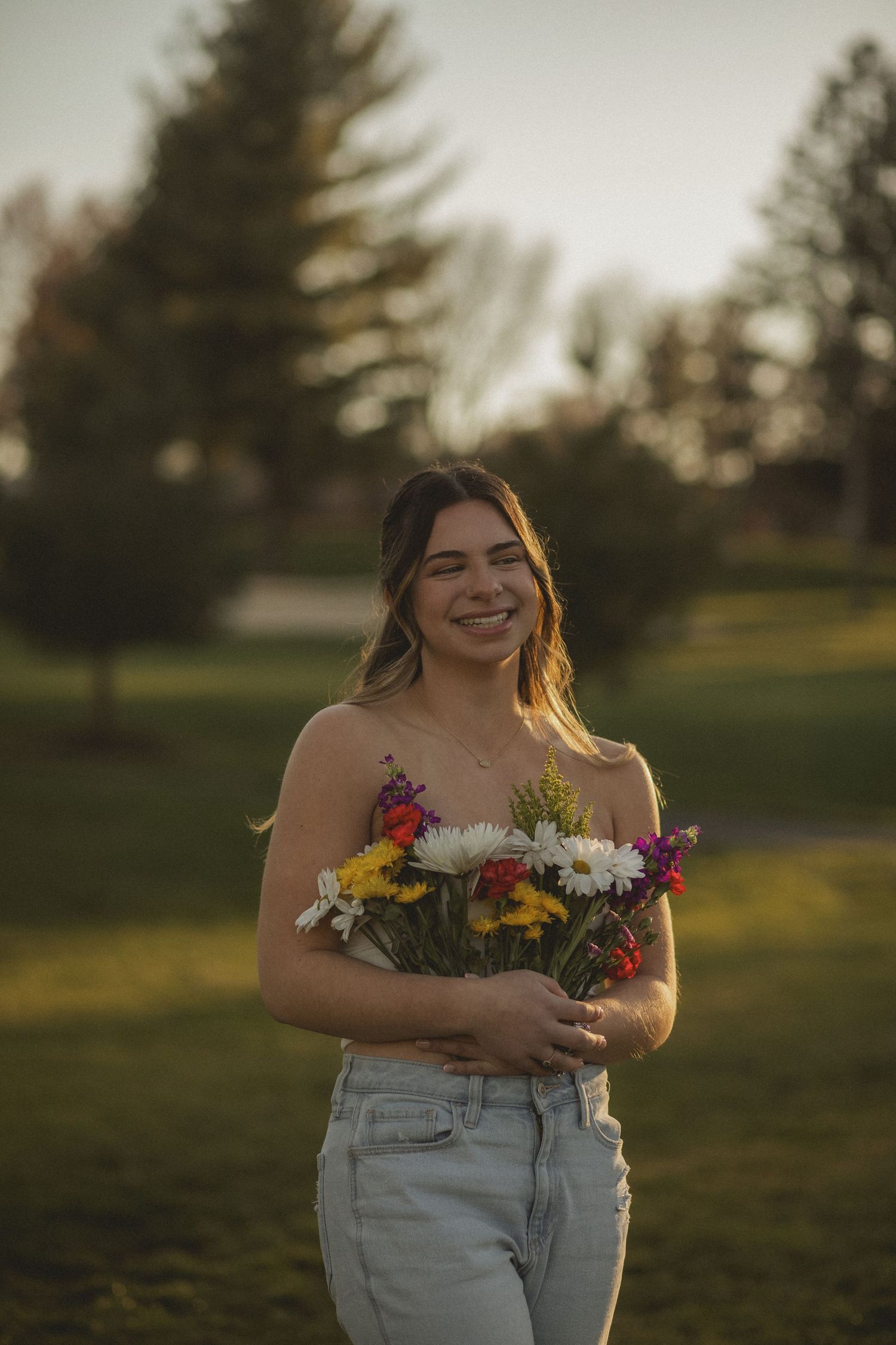 A woman smiles warmly, holding a bouquet of colorful flowers, bathed in the golden glow of the setting sun.