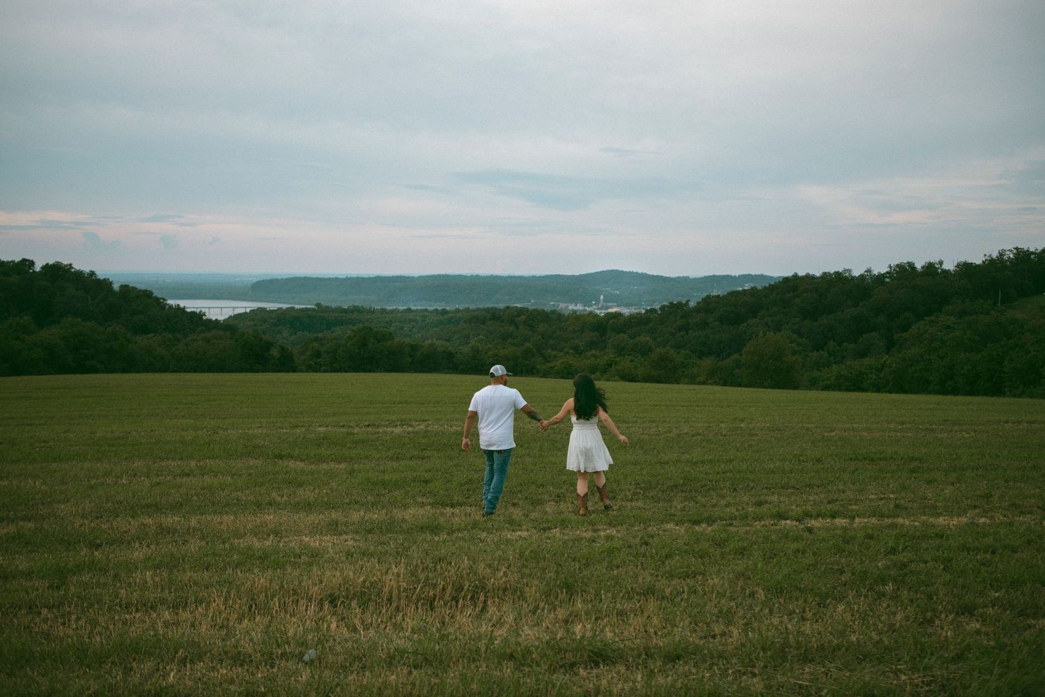 A couple run hand in hand across a vast grassy field with mountains and water visible in the distance in a dreamy couples session.
