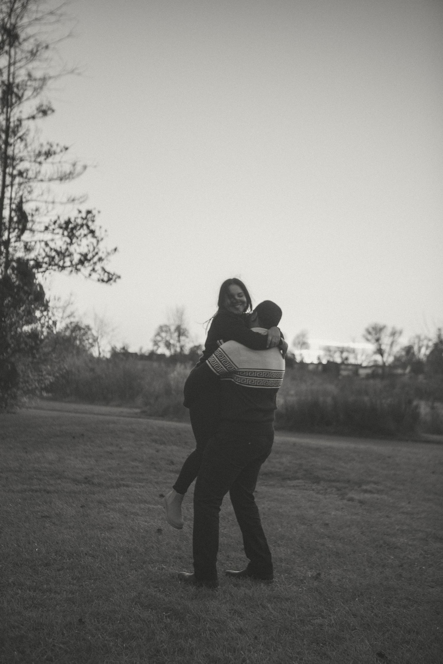 Black-and-white photo of a couple in a joyful embrace outdoors as the man lifts the woman, capturing a candid and romantic moment at sunset.