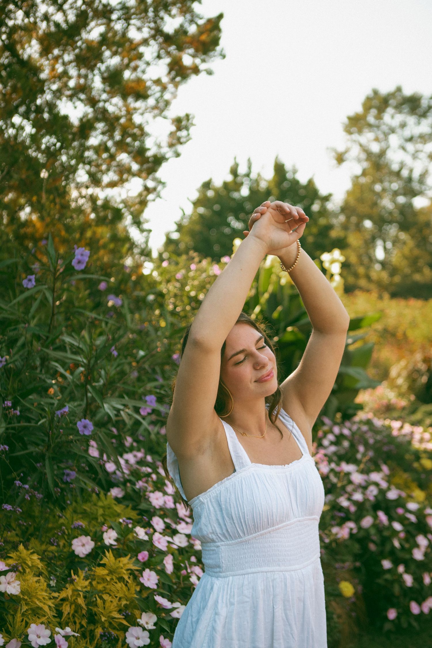 Young woman in a white dress enjoys a peaceful moment in a blooming garden, basking in the warm sunlight with her arms raised in relaxation.