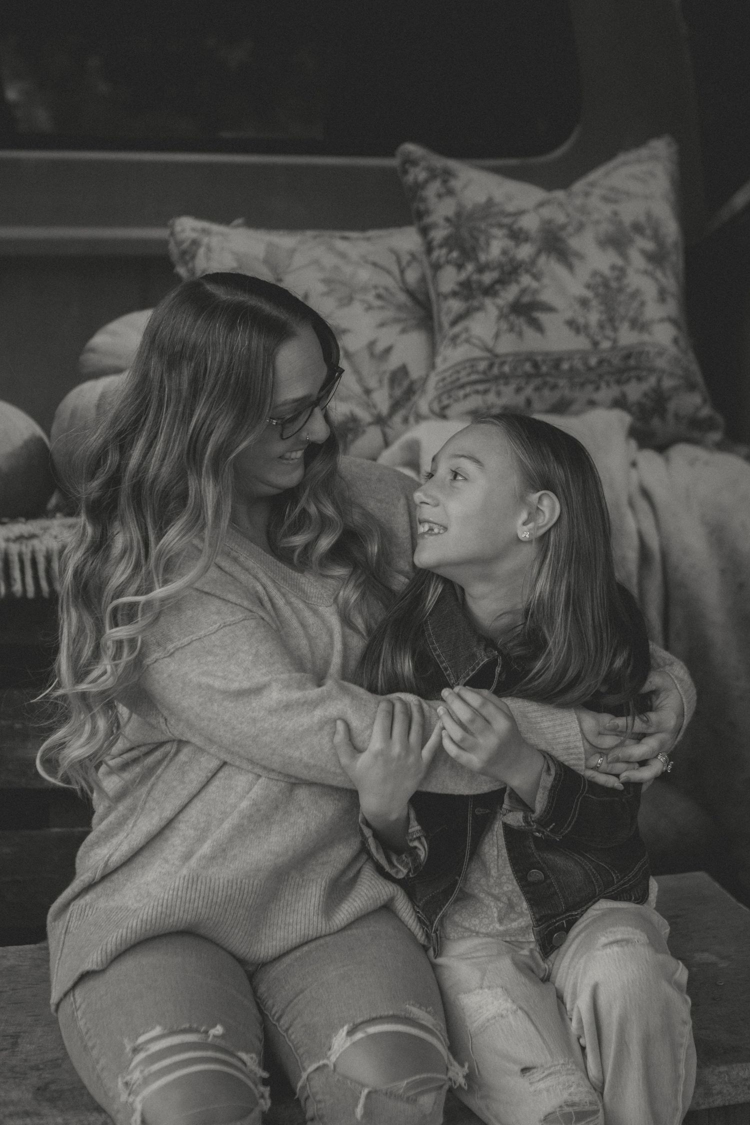 A mother and daughter share a warm embrace, smiling at each other in a cozy autumn setting with pumpkins and soft pillows in the background.