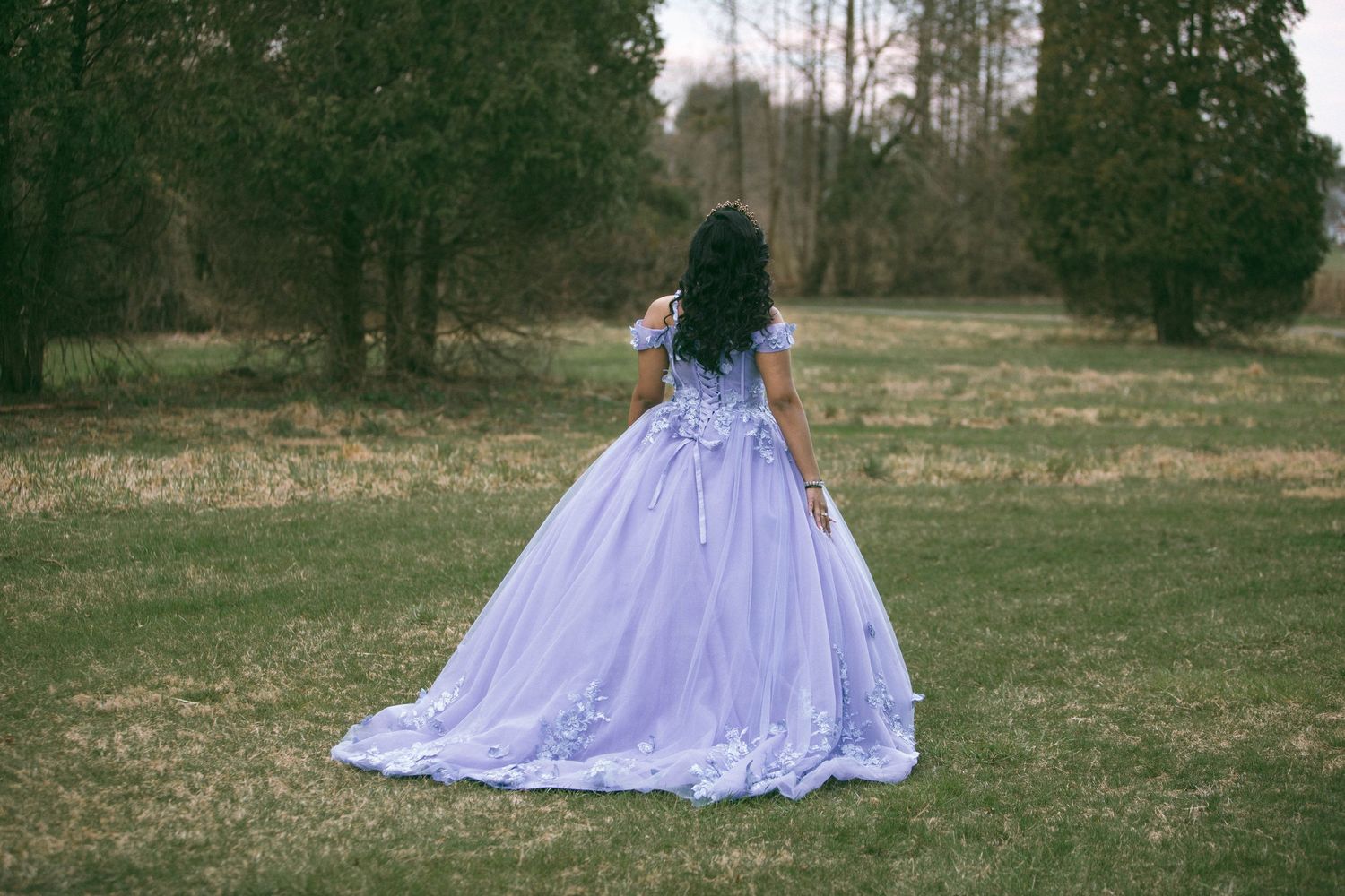 Quinceañera in lavender ball gown stands in a meadow with trees in the background by D'Gallery Photography in Lancaster, PA.
