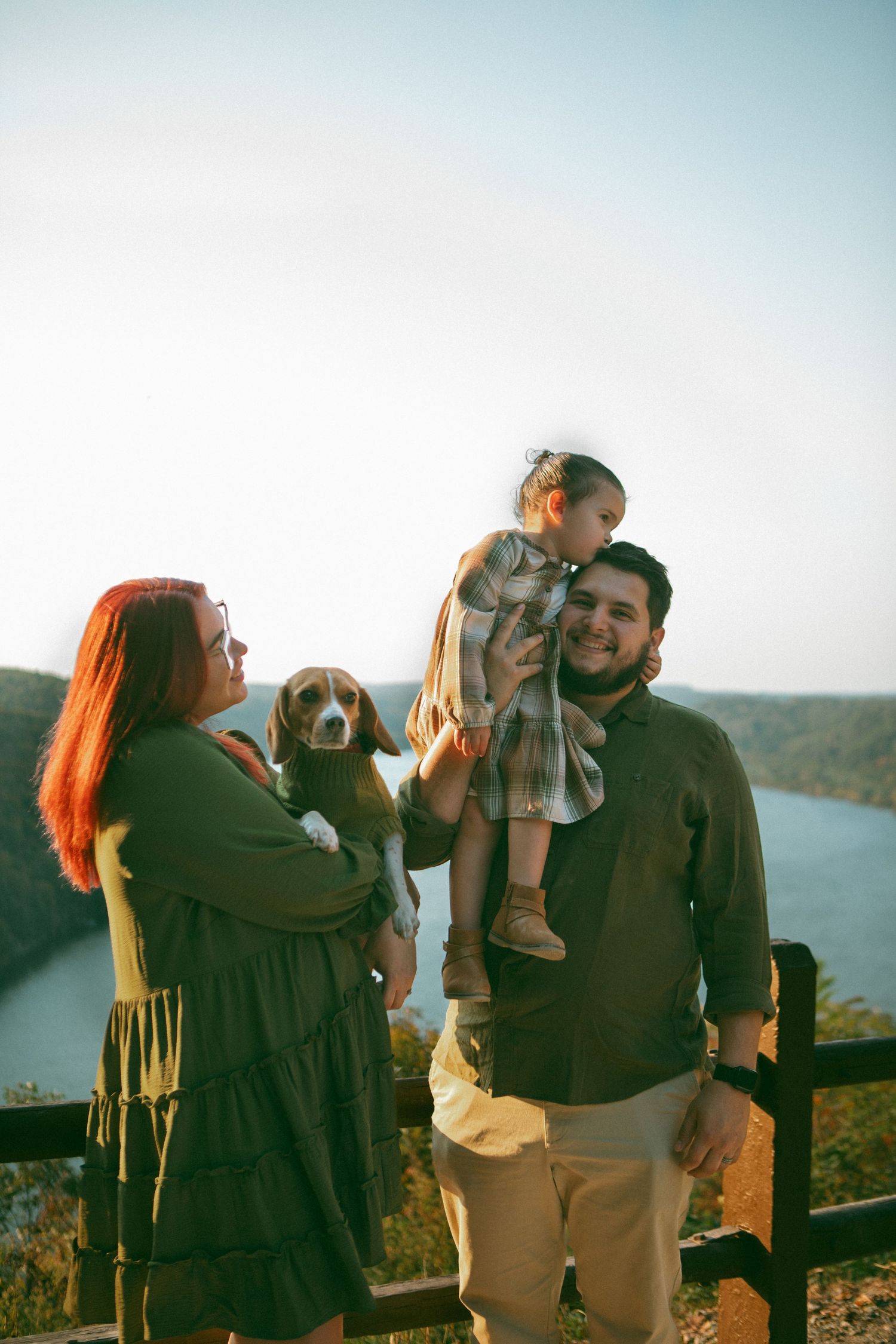 A loving family shares a warm moment at a scenic overlook, with a child kissing her father and a cozy pup in the mother's arms.