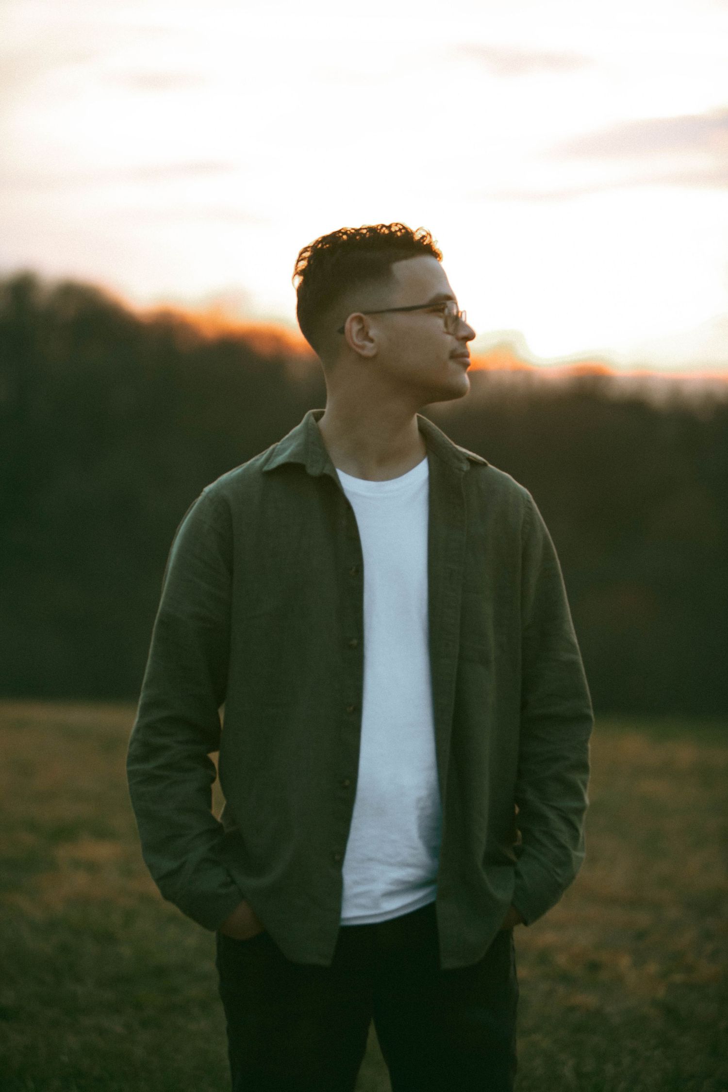A man from a couples session in Wrightsville, PA, looks away into the distance in a grassy field at sunset with trees in background.