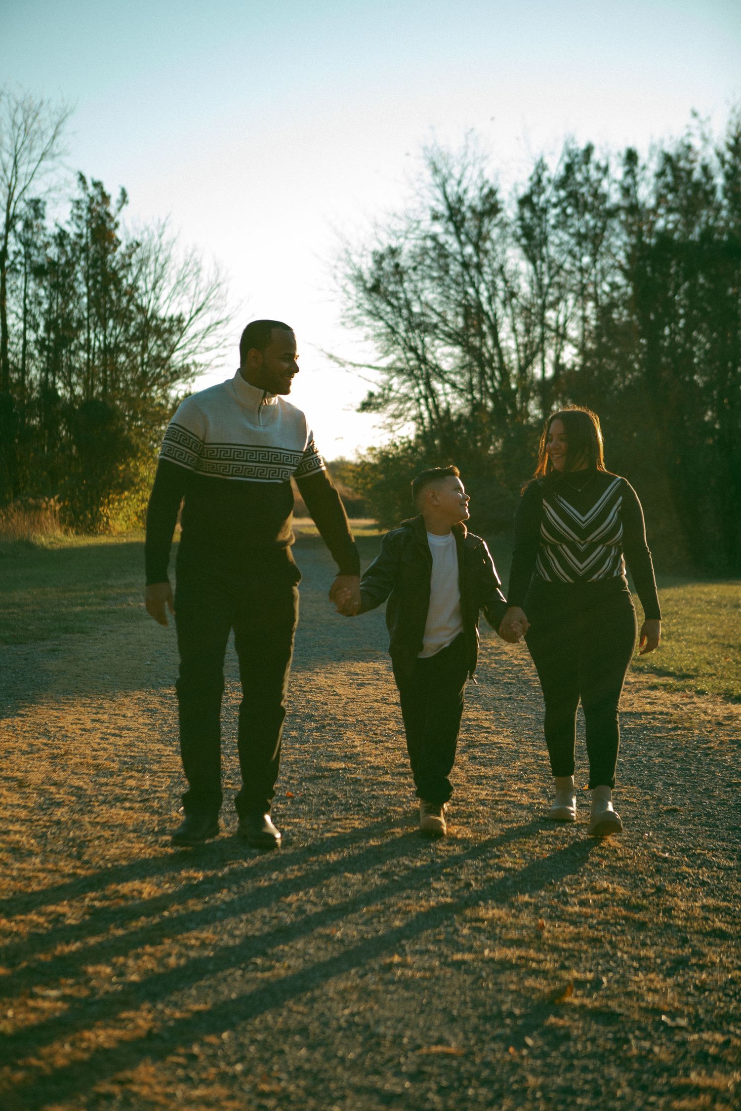 Happy family walking hand in hand on a sunlit path, sharing smiles and love in a golden-hour setting with autumn trees in the background.