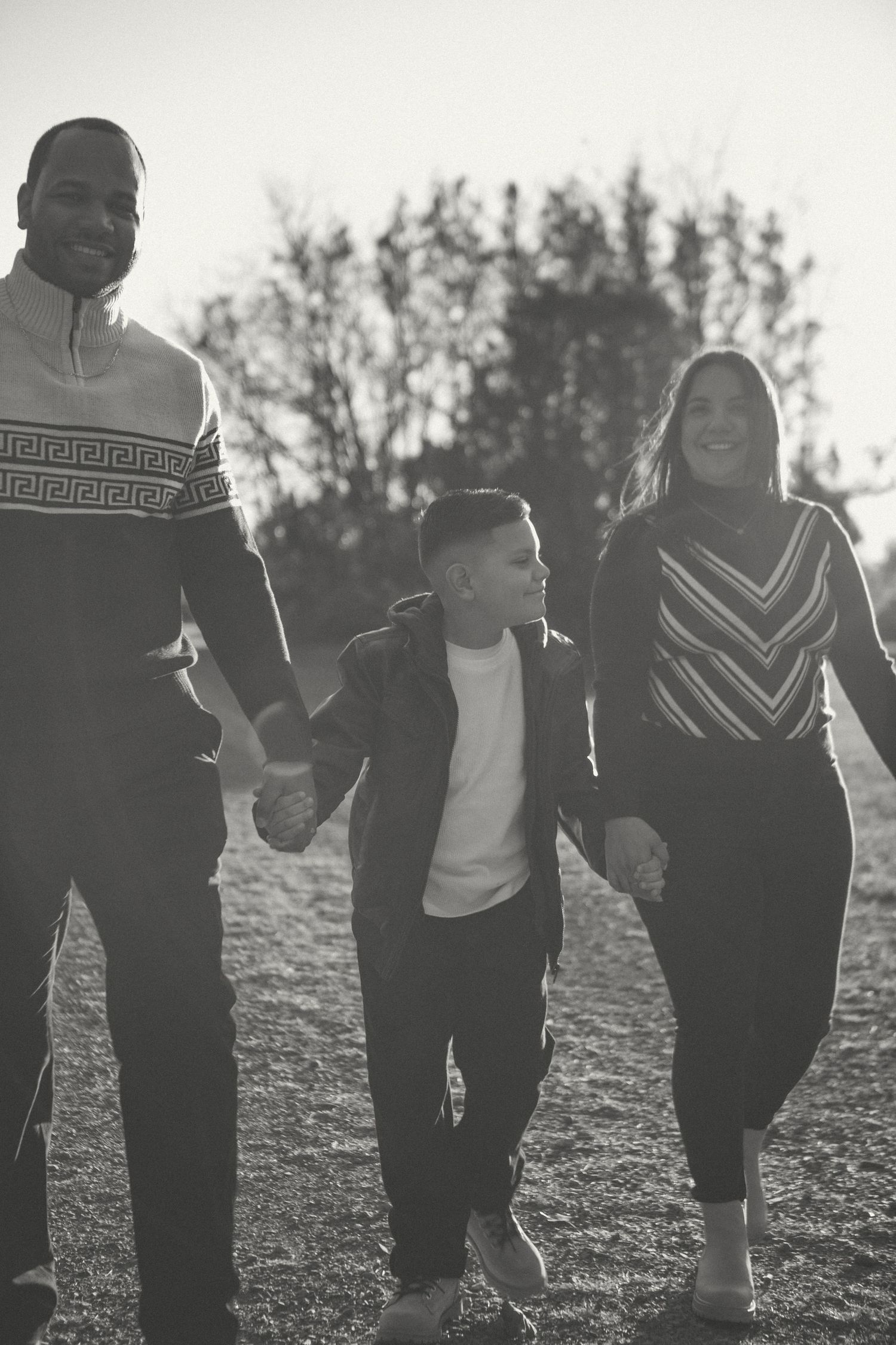 Happy family walking hand in hand outdoors surrounded by nature at golden hour, smiling together in a black-and-white candid shot.