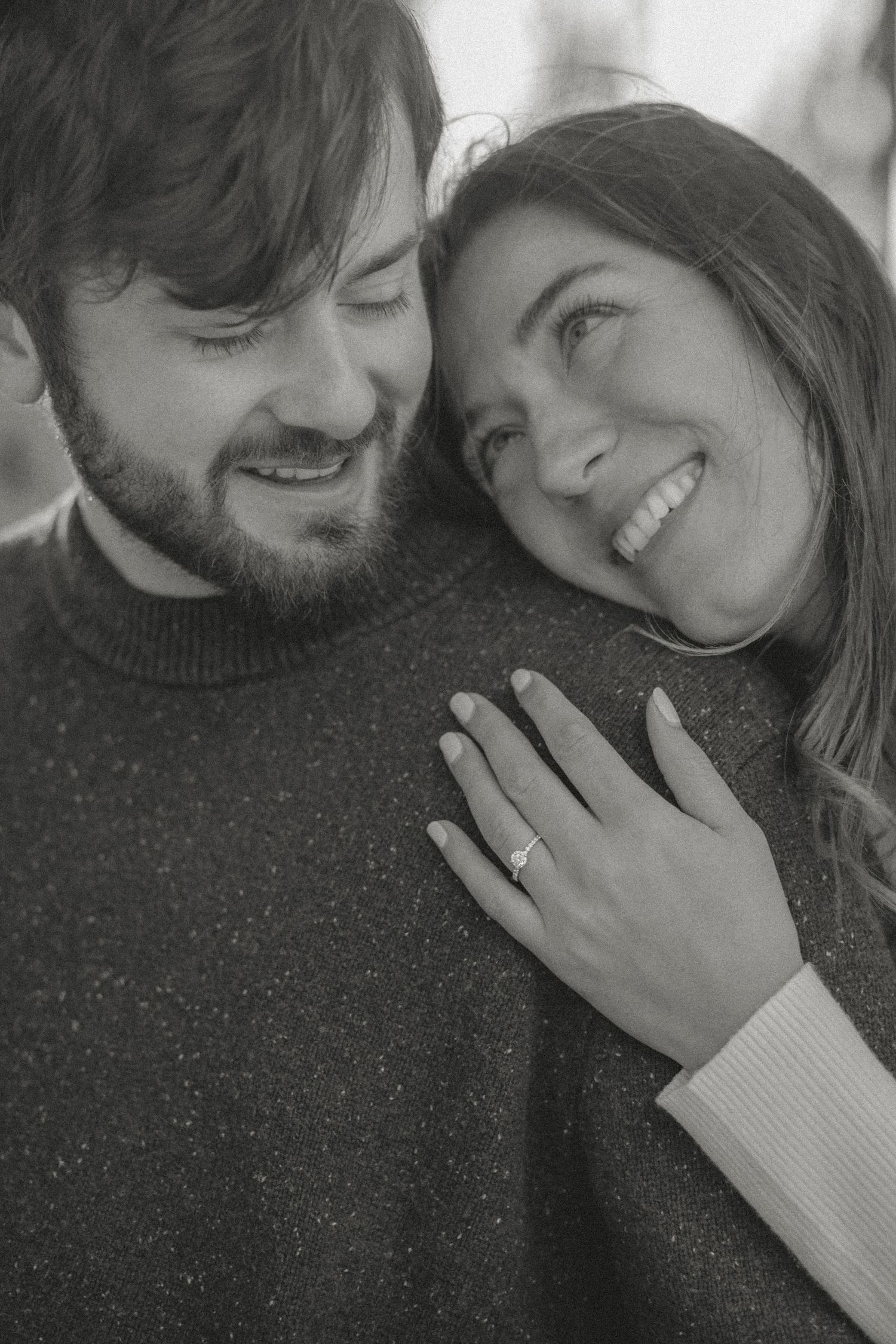 Black-and-white engagement photo of a joyful couple, with the woman leaning on her partner’s shoulder, showcasing her elegant diamond ring.