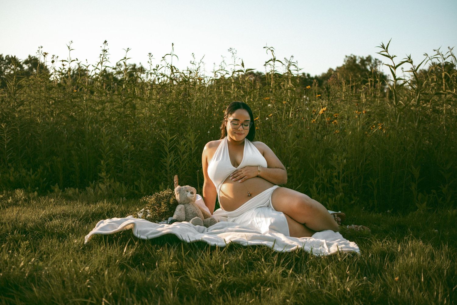 A maternity session with a pregnant woman sitting in a grassy field at golden hour with tall wildflowers in the background.
