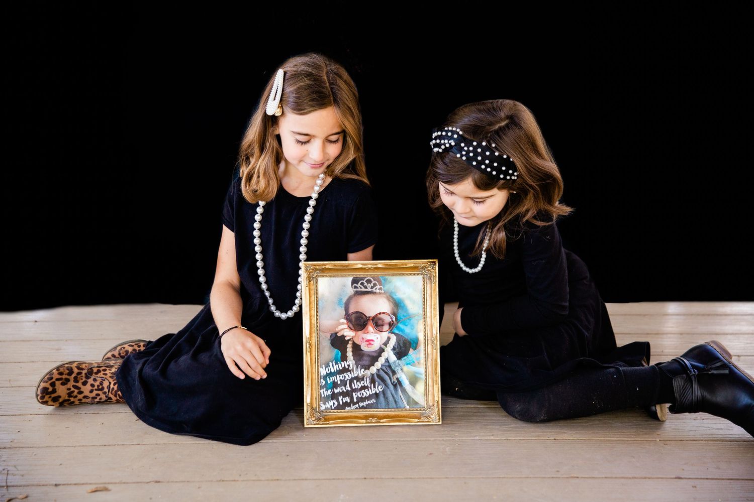 Two children in vintage-style black dresses sit beside a framed photograph on a wooden floor.