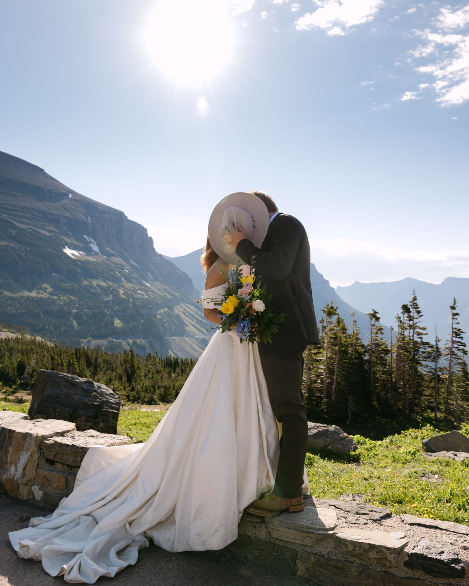 A couple in wedding attire shares an intimate moment against a backdrop of majestic mountain peaks and evergreen forest.