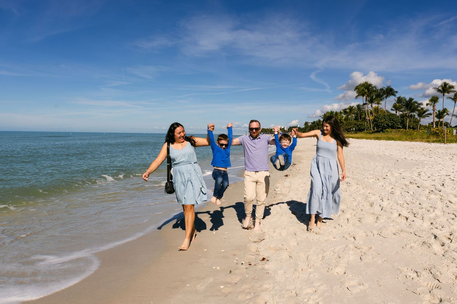 A group in coordinated blue and gray outfits walks joyfully along a pristine beach shoreline on a sunny day.