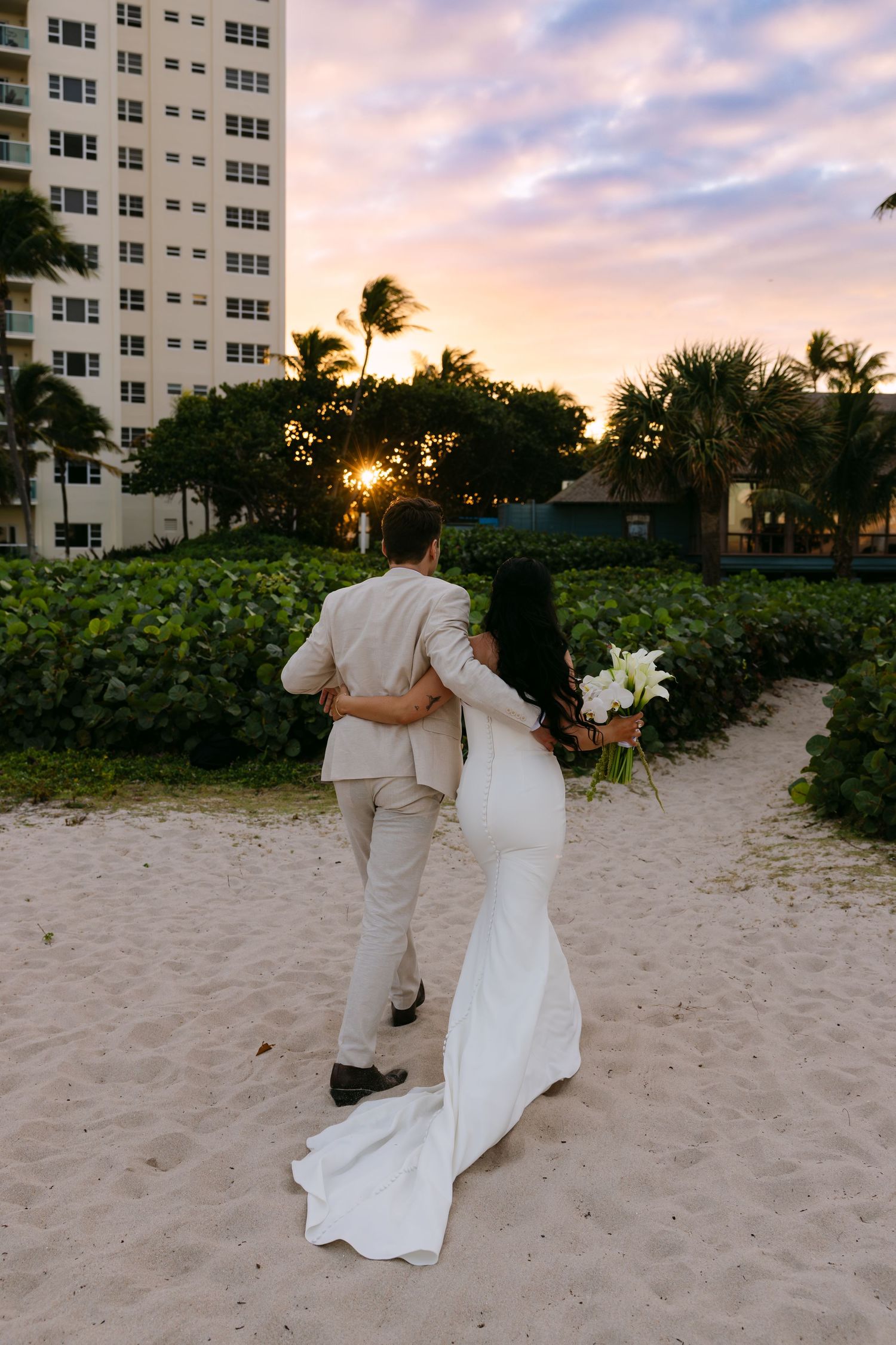 A couple in white attire embrace while walking on a sandy beach at sunset with palm trees and buildings in the background.