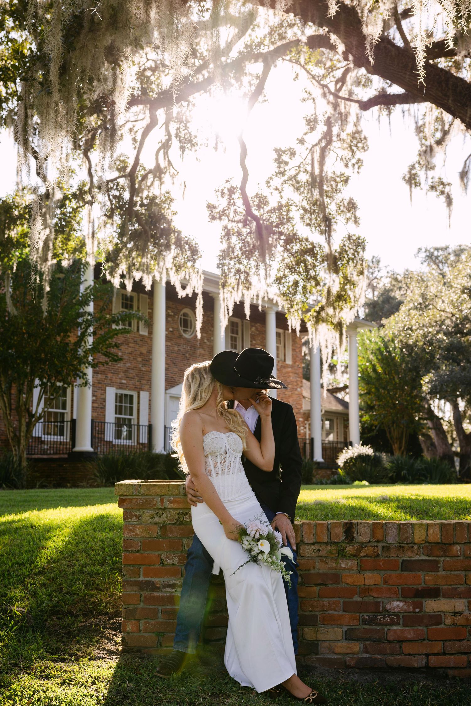 Romantic couple embraces by a rustic brick wall under Spanish moss draped trees at a Southern plantation estate.