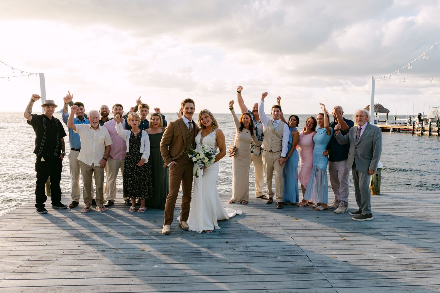 Wedding party celebrating on a waterfront dock at sunset, bride and groom centered among cheering guests.