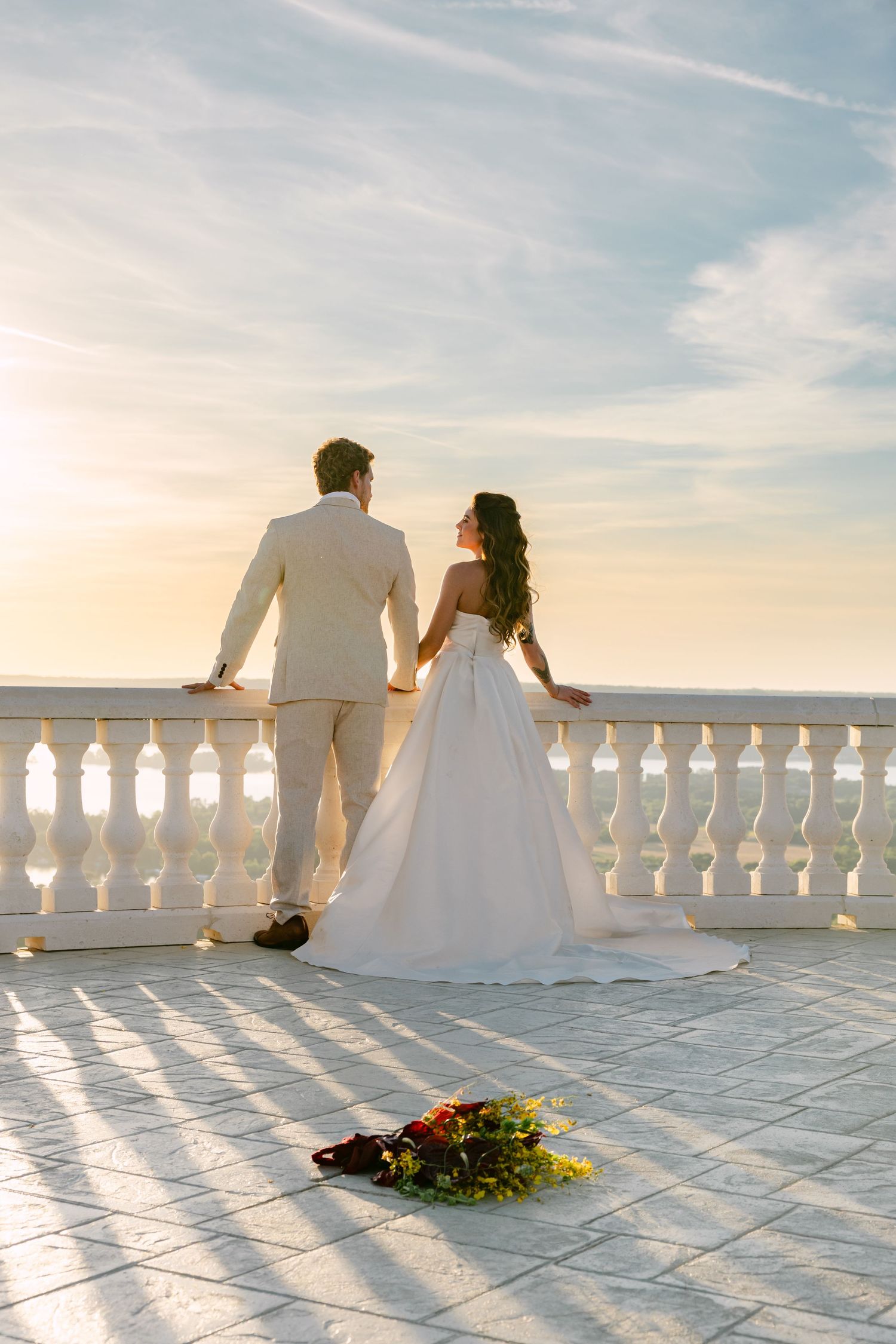 Bride and groom stand at a white balustrade terrace overlooking a golden sunset, with a floral bouquet on the floor.