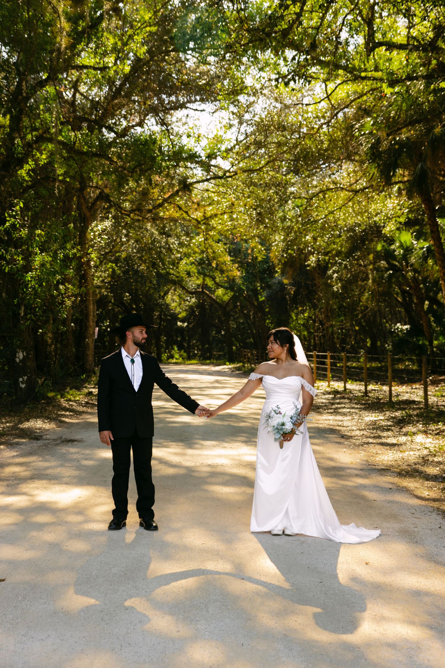 Bride and groom hold hands on a sunlit path surrounded by lush green trees in an outdoor wedding photo.