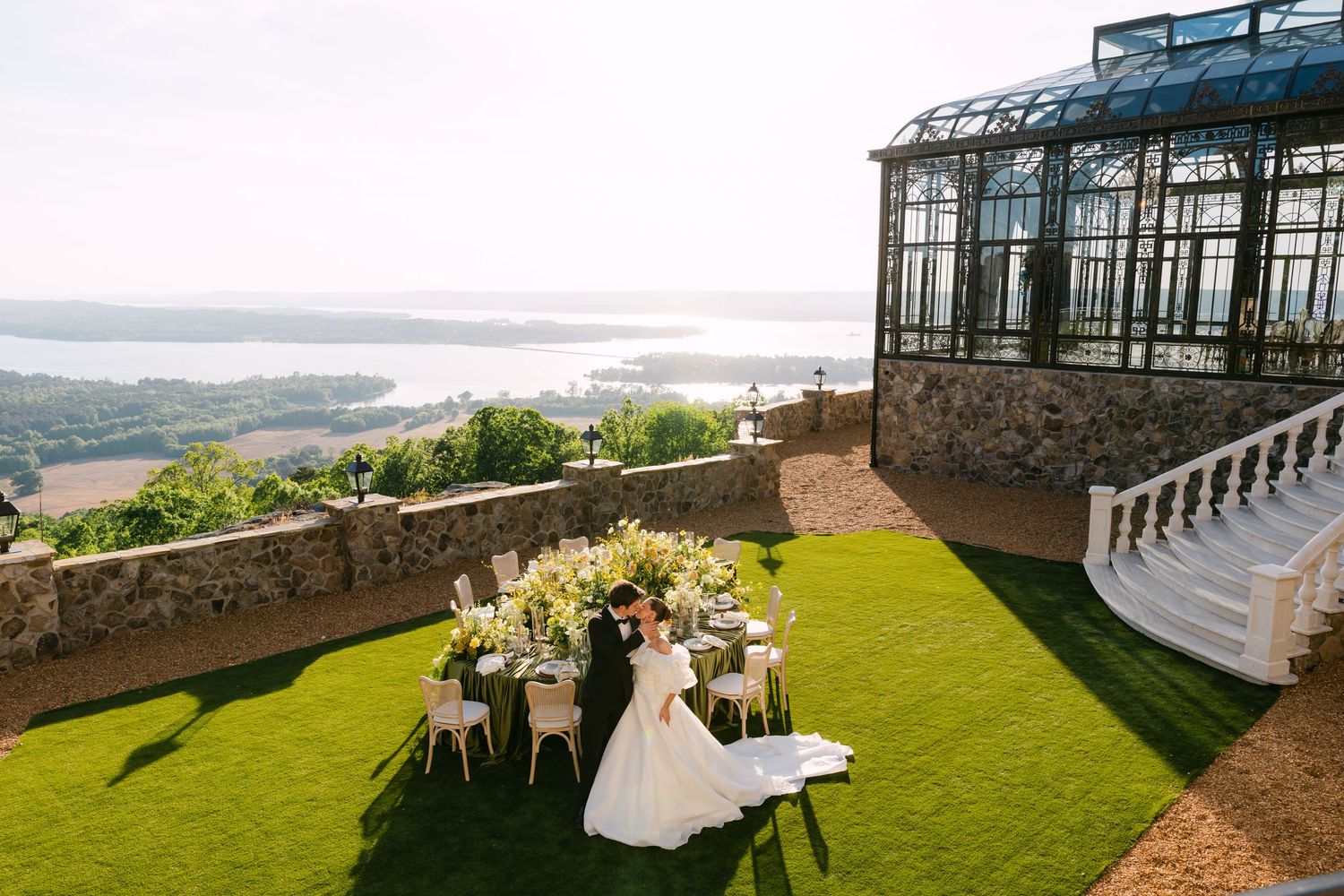 Bride and groom stand at floral wedding table on lush green terrace overlooking scenic river valley with glass greenhouse.
