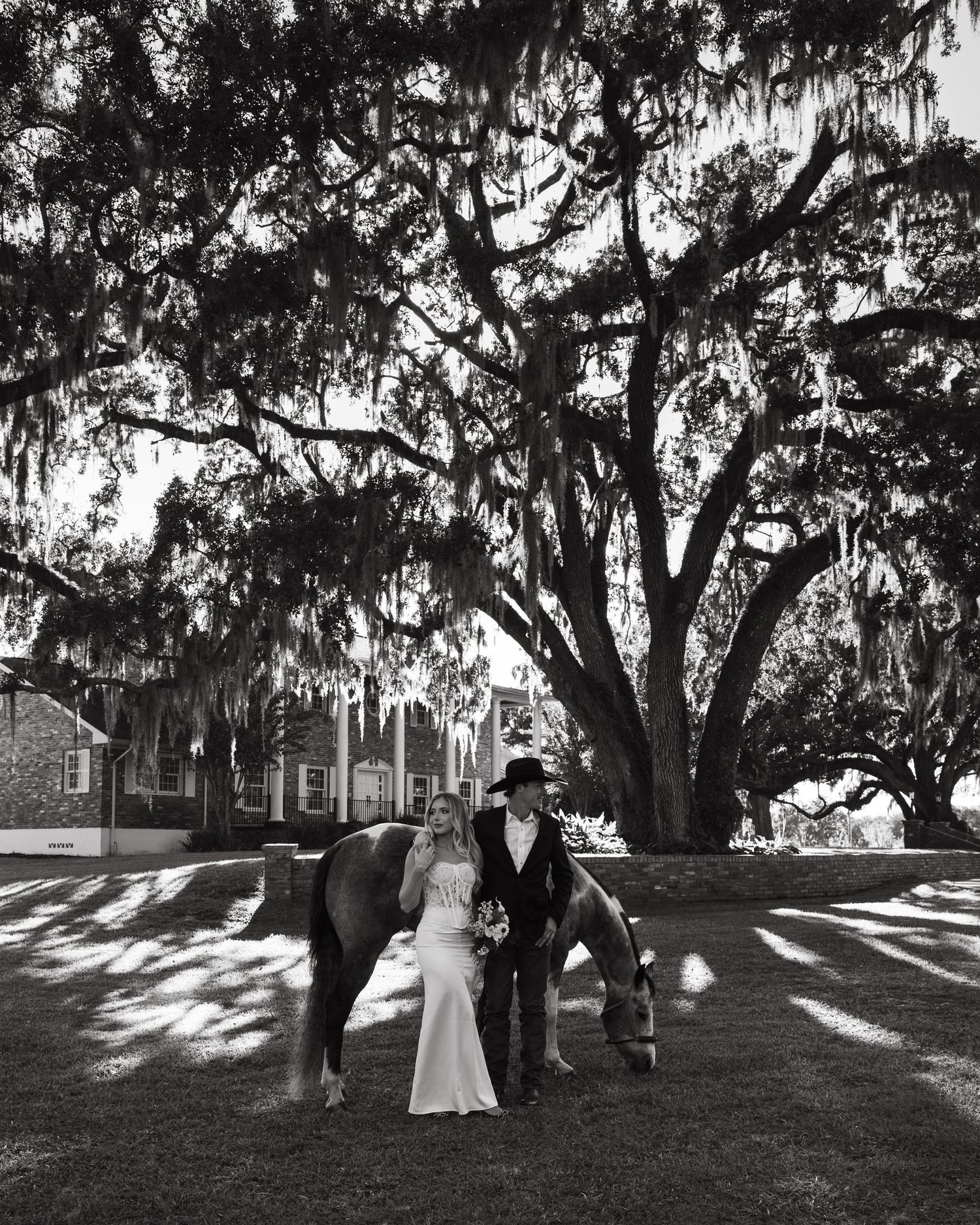A black and white photograph captures a romantic moment under moss-draped oak trees on a sunlit lawn.