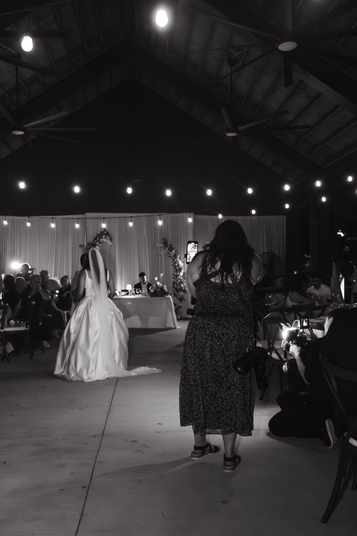 A wedding reception scene in a dimly lit venue with string lights overhead and silhouettes of guests dancing.