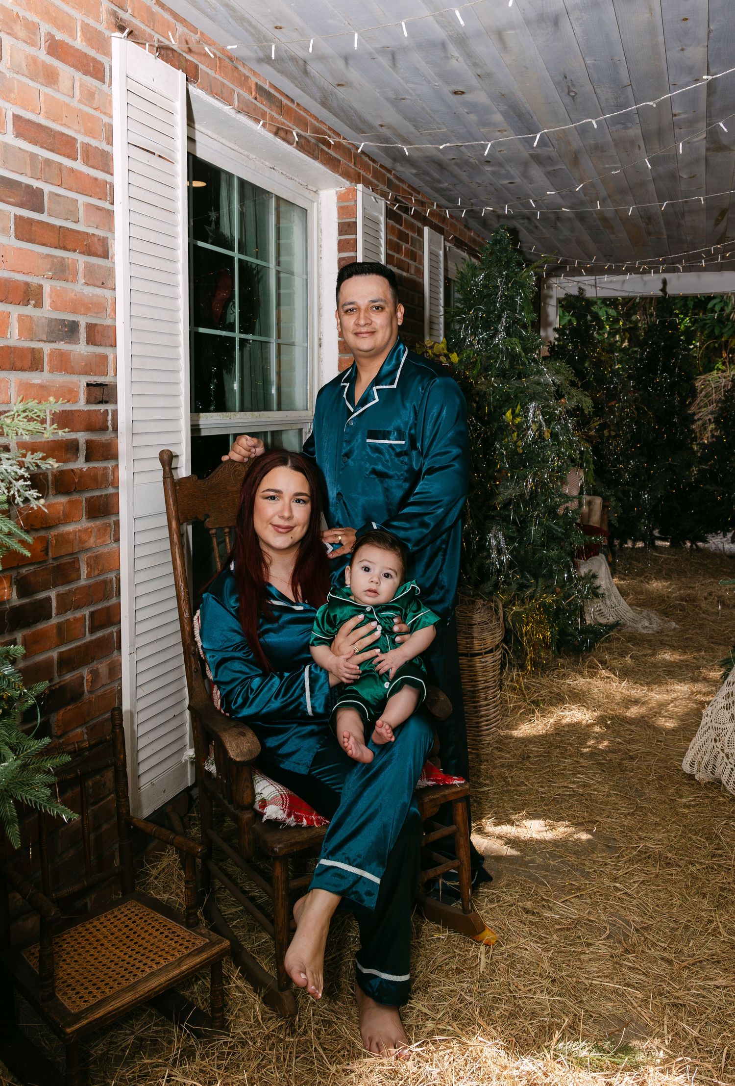 A family wearing matching blue satin pajamas poses together in a rustic indoor setting with brick walls and twinkle lights.