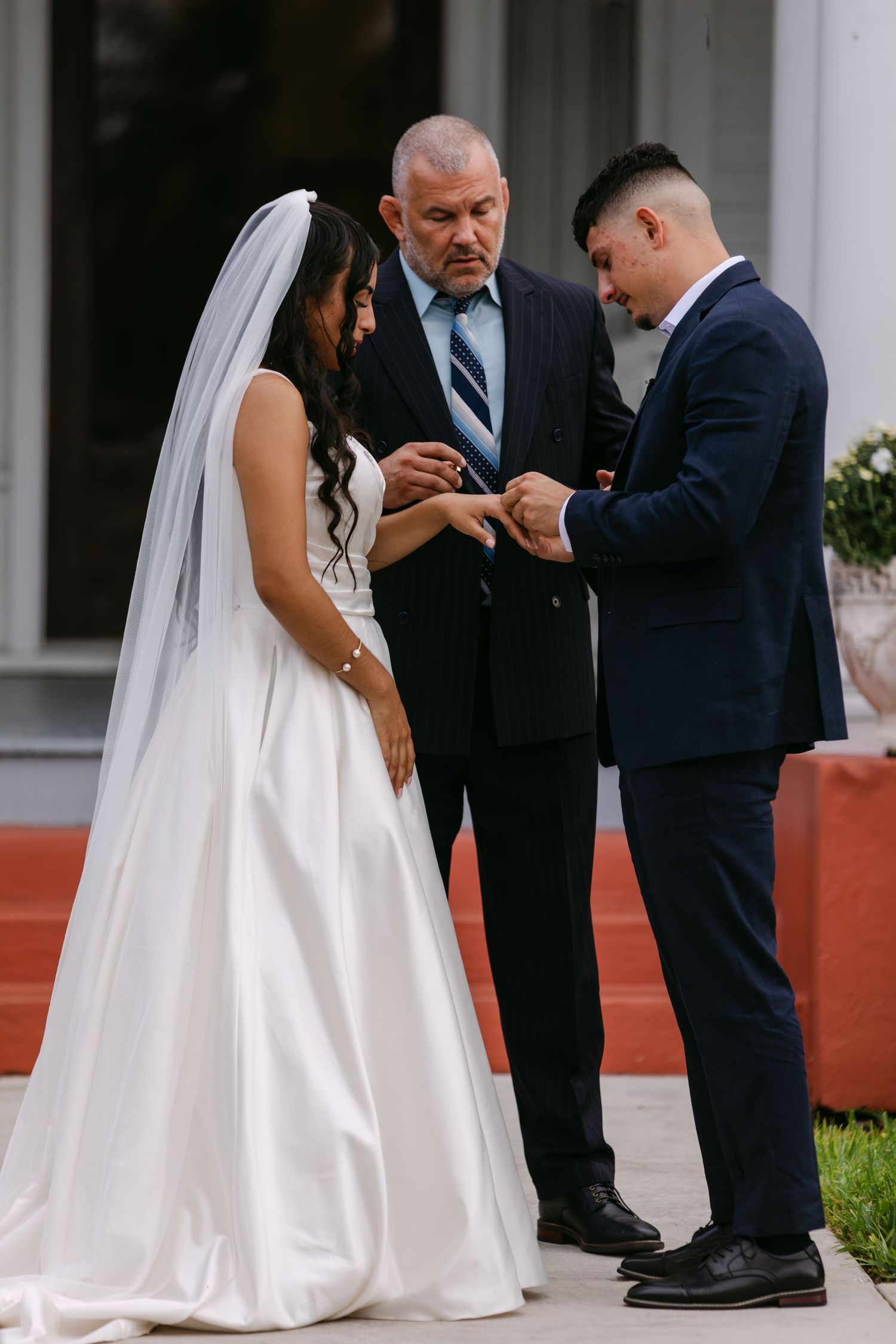 Groom placing wedding ring on bride's finger during outdoor ceremony, officiant watching, bride in white gown and veil.