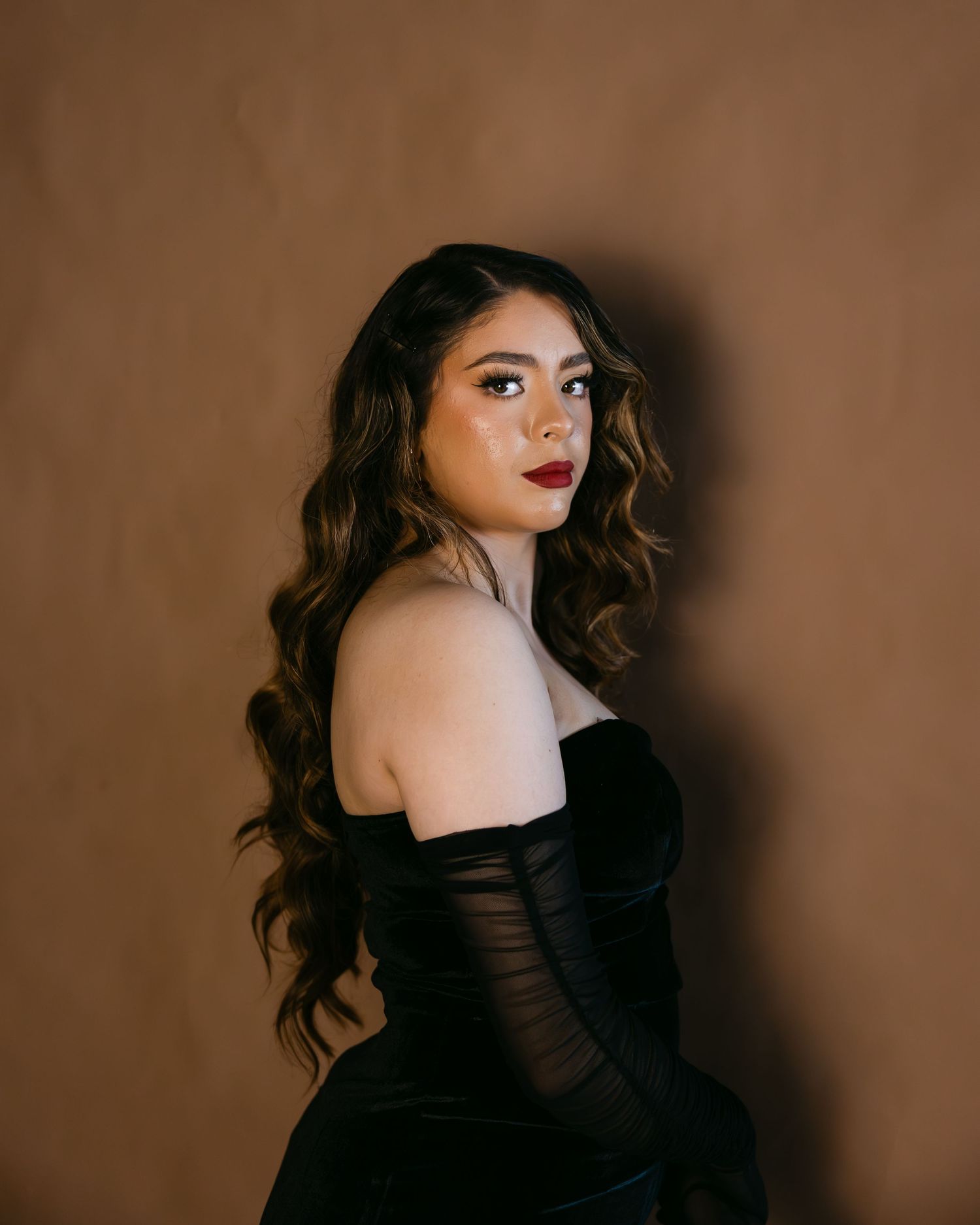 Portrait against brown backdrop of person in black off-shoulder dress with long wavy dark hair and red lipstick.