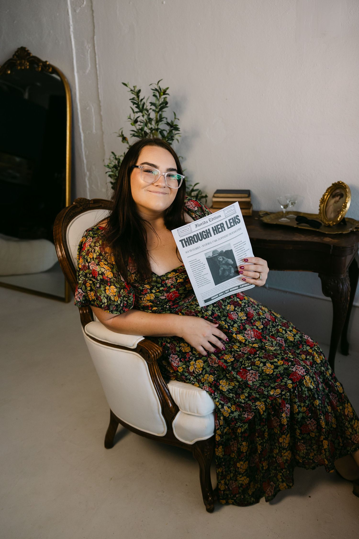 A person in a floral dress lounges in a white chair while holding and reading a book.