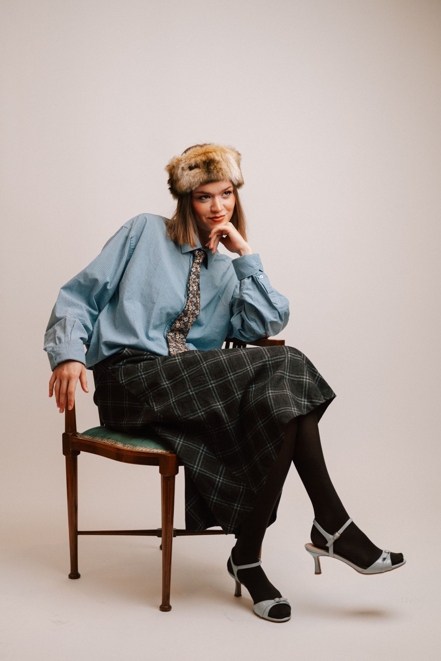 A person in a light blue shirt and plaid pants sits casually on a wooden chair against a neutral background.