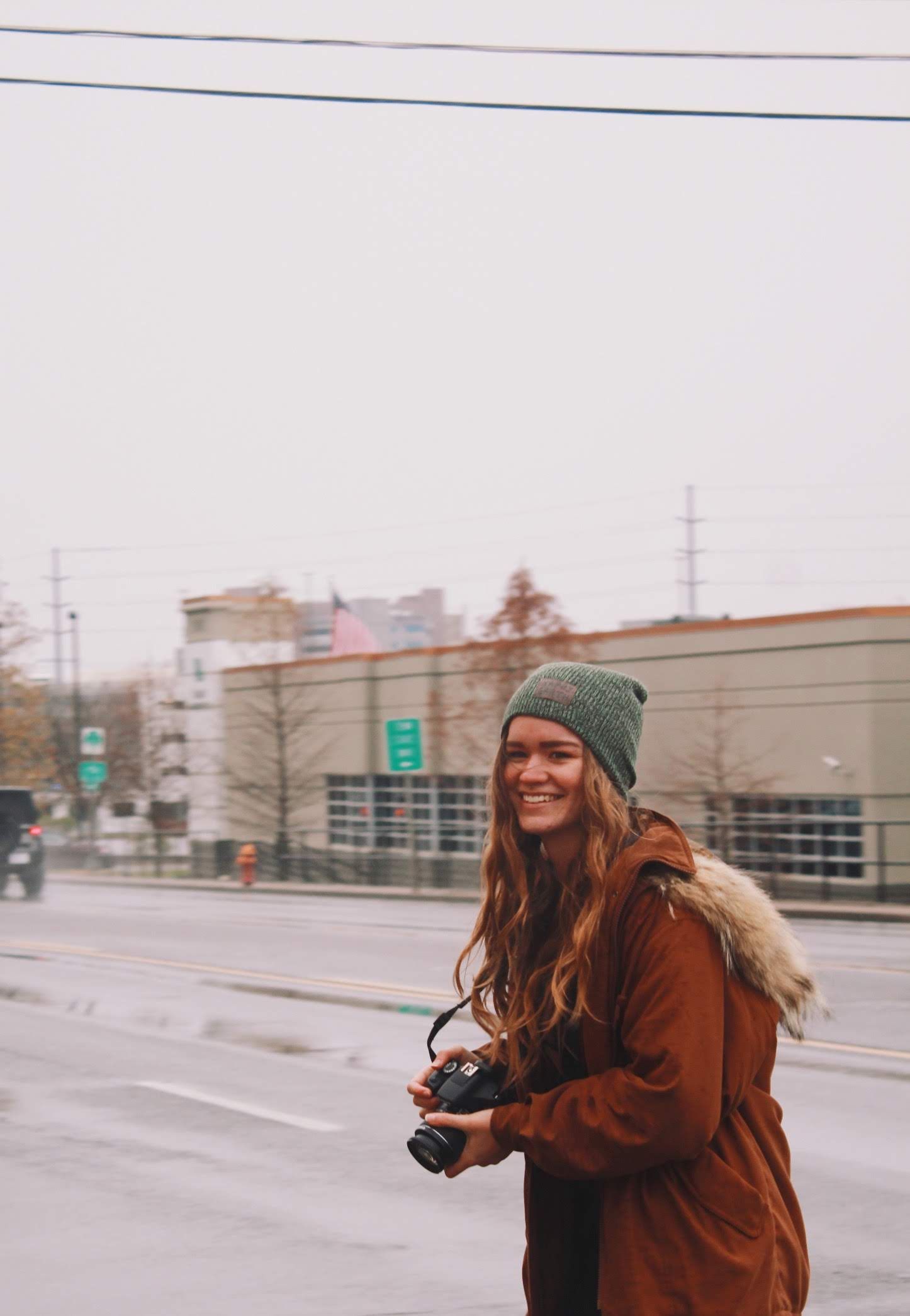 Person in brown coat and green beanie taking photos outdoors on a cloudy winter day.