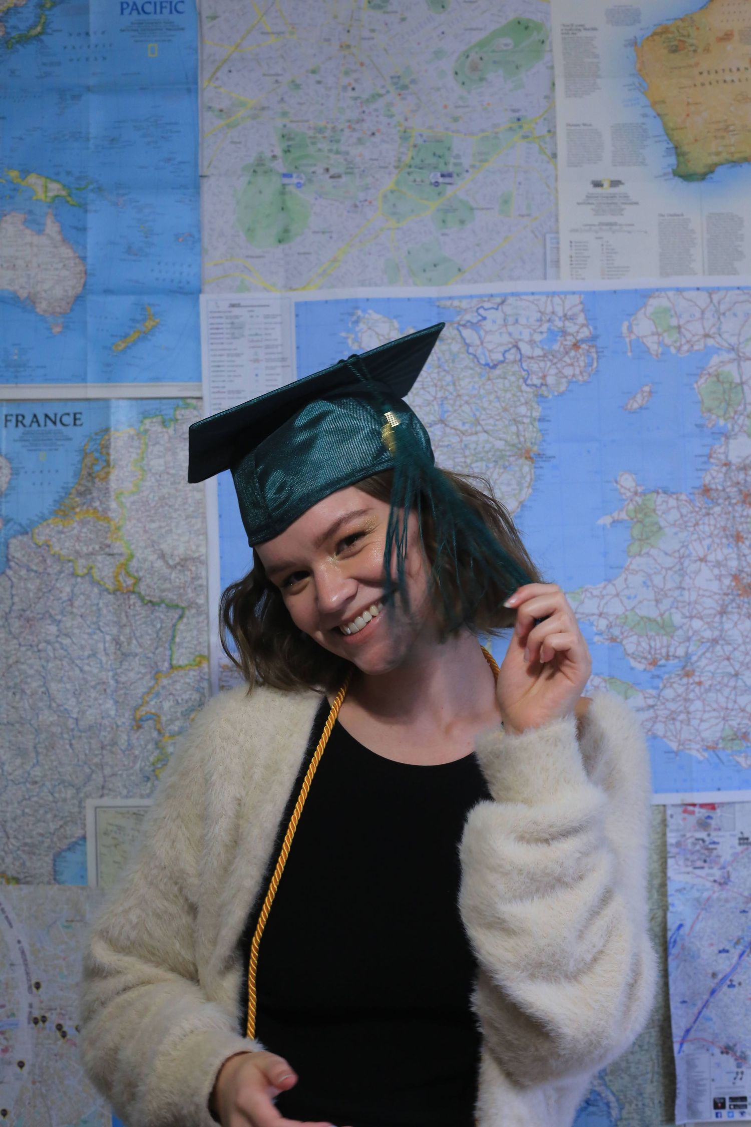 A graduate in a black cap and gown with a gold tassel poses against a wall of world maps.