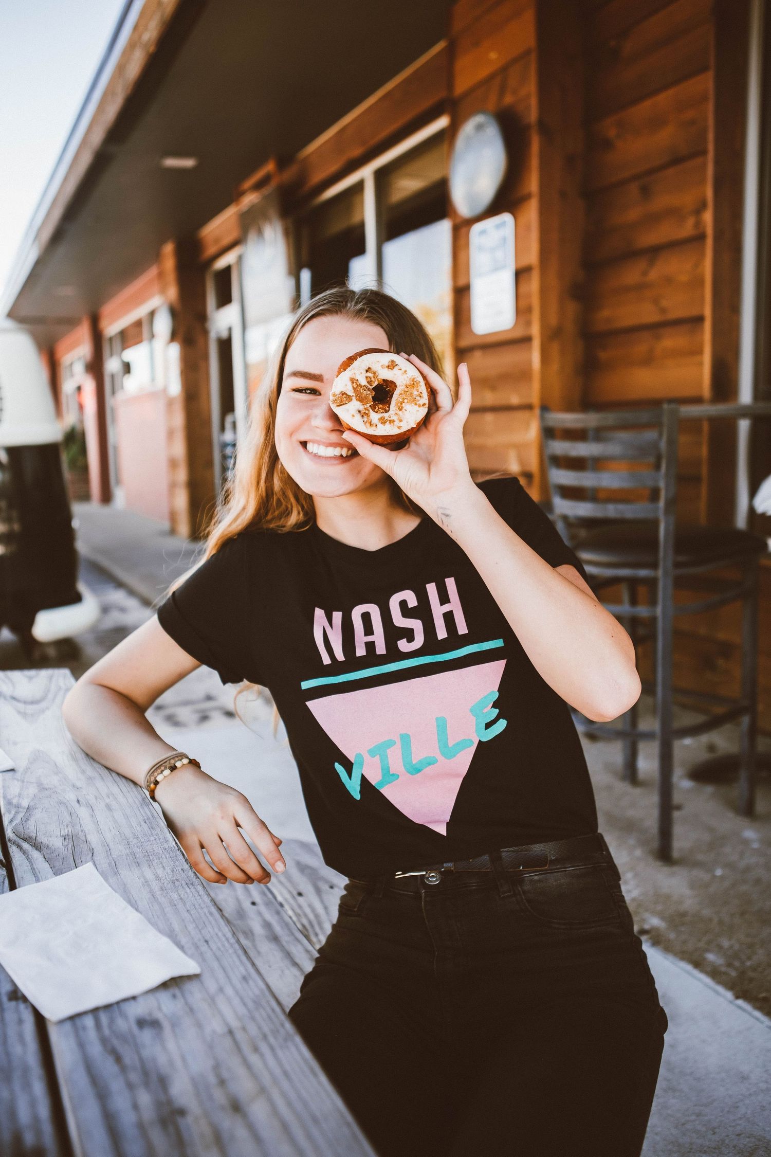 Someone wearing a black Nashville t-shirt holds a donut up to their eye while smiling outside a rustic wooden building.
