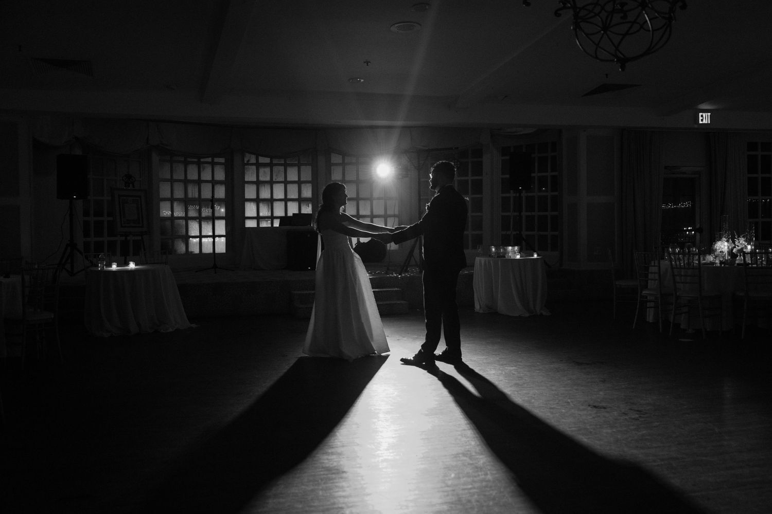 A silhouetted couple shares their first dance in a dimly lit ballroom with dramatic backlighting creating long shadows.