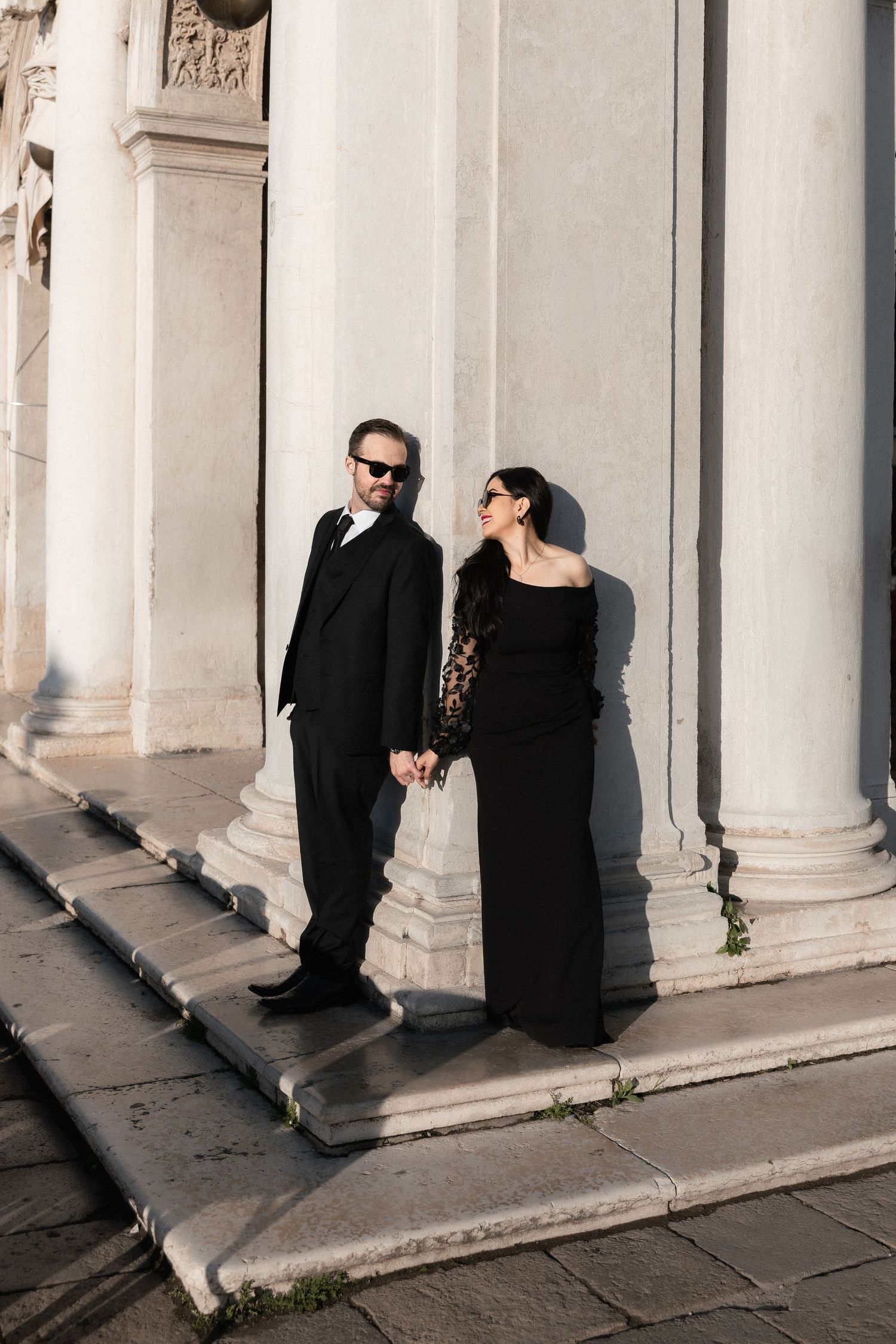 A couple in formal black attire poses on white stone steps against classical columns in dramatic lighting.