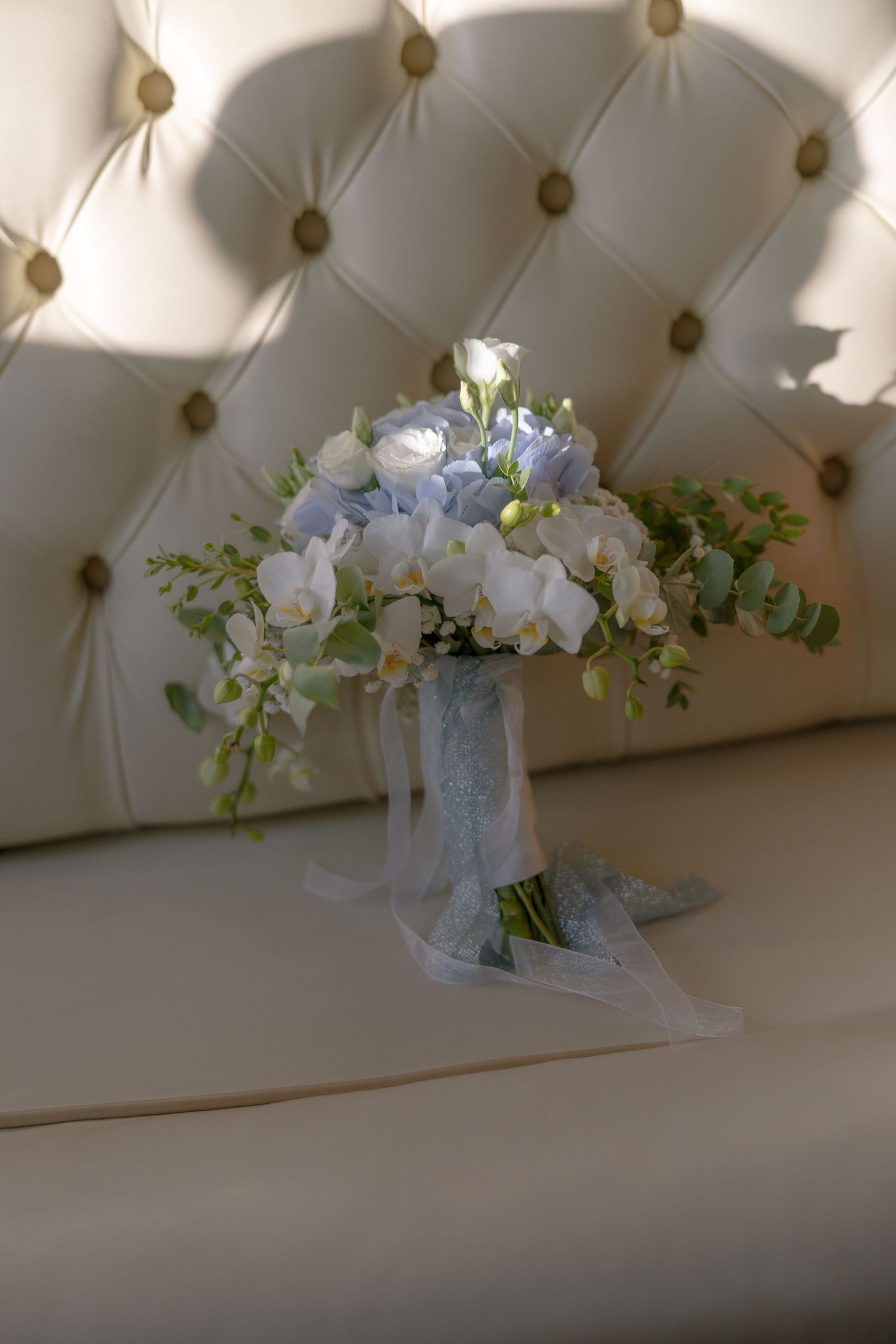 A delicate bridal bouquet with white and light blue flowers tied with lace ribbon sits against a tufted cream backdrop.