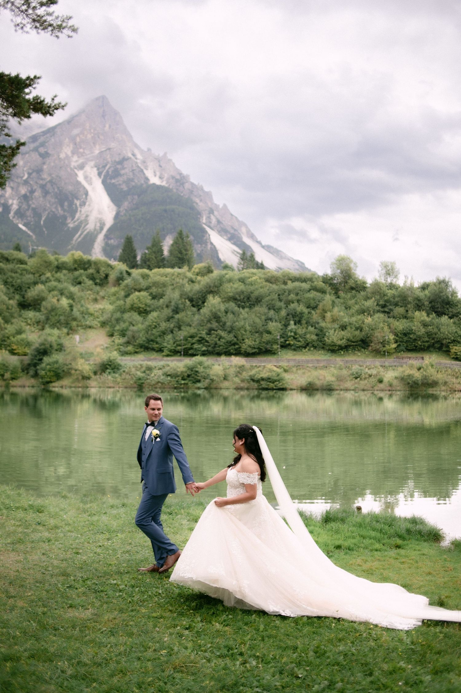 Bride and groom hold hands by a mountain lake, she in a flowing white gown with veil, he in a blue suit.