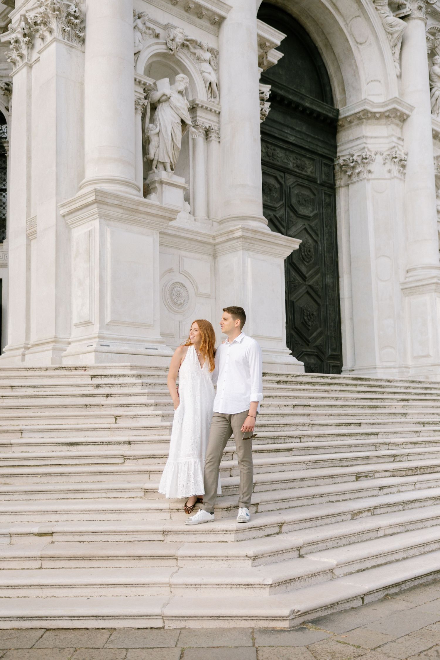 A couple stands on ornate white stone steps of a grand cathedral with intricate baroque architectural details.