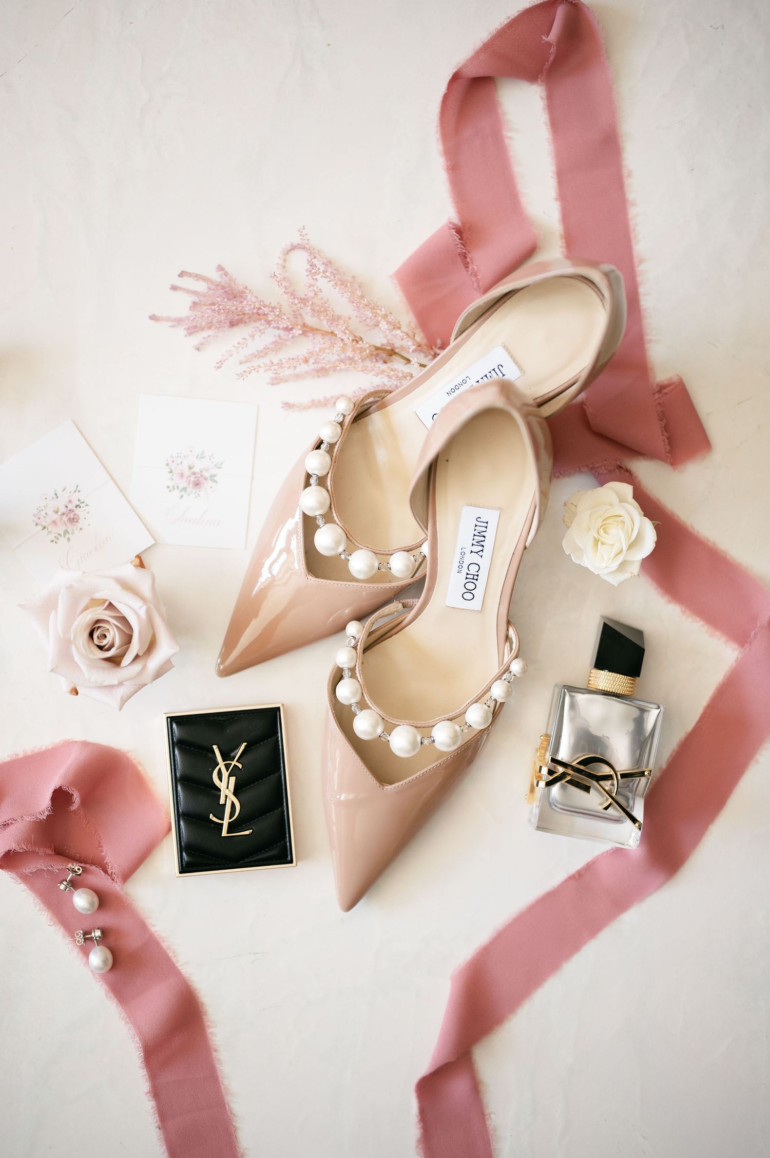 Elegant pink accessories and YSL makeup arranged with blush pink ribbons and roses in a feminine wedding flat lay. Jimmy Choo shoes
