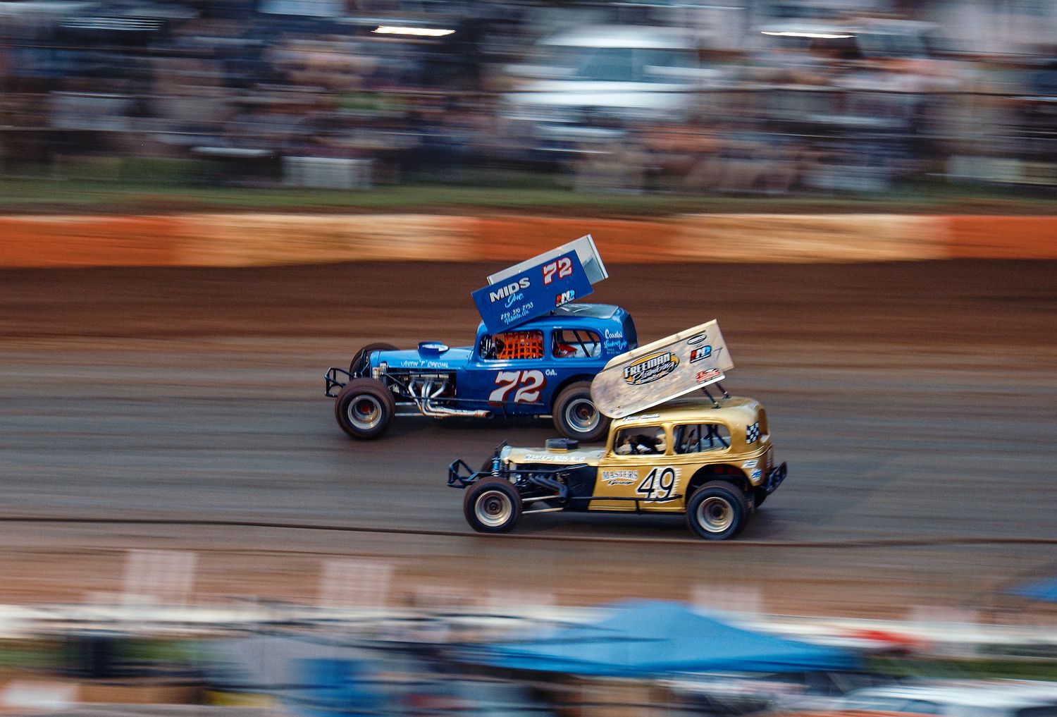 Two vintage race cars speeding past on a dirt track with motion blur creating dynamic racing action.