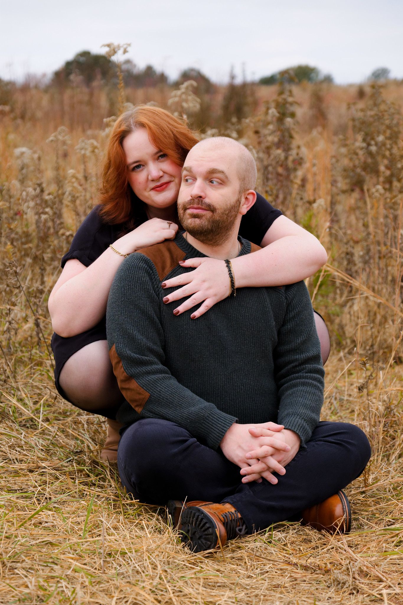 A couple embraces while sitting in a golden autumn field with tall dried grasses and plants surrounding them.