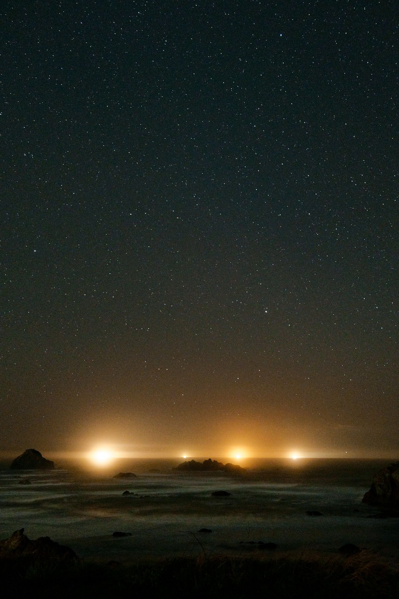 A starry night sky over the pacific ocean. Multiple lights from a fishing boat can be seen on the horizon