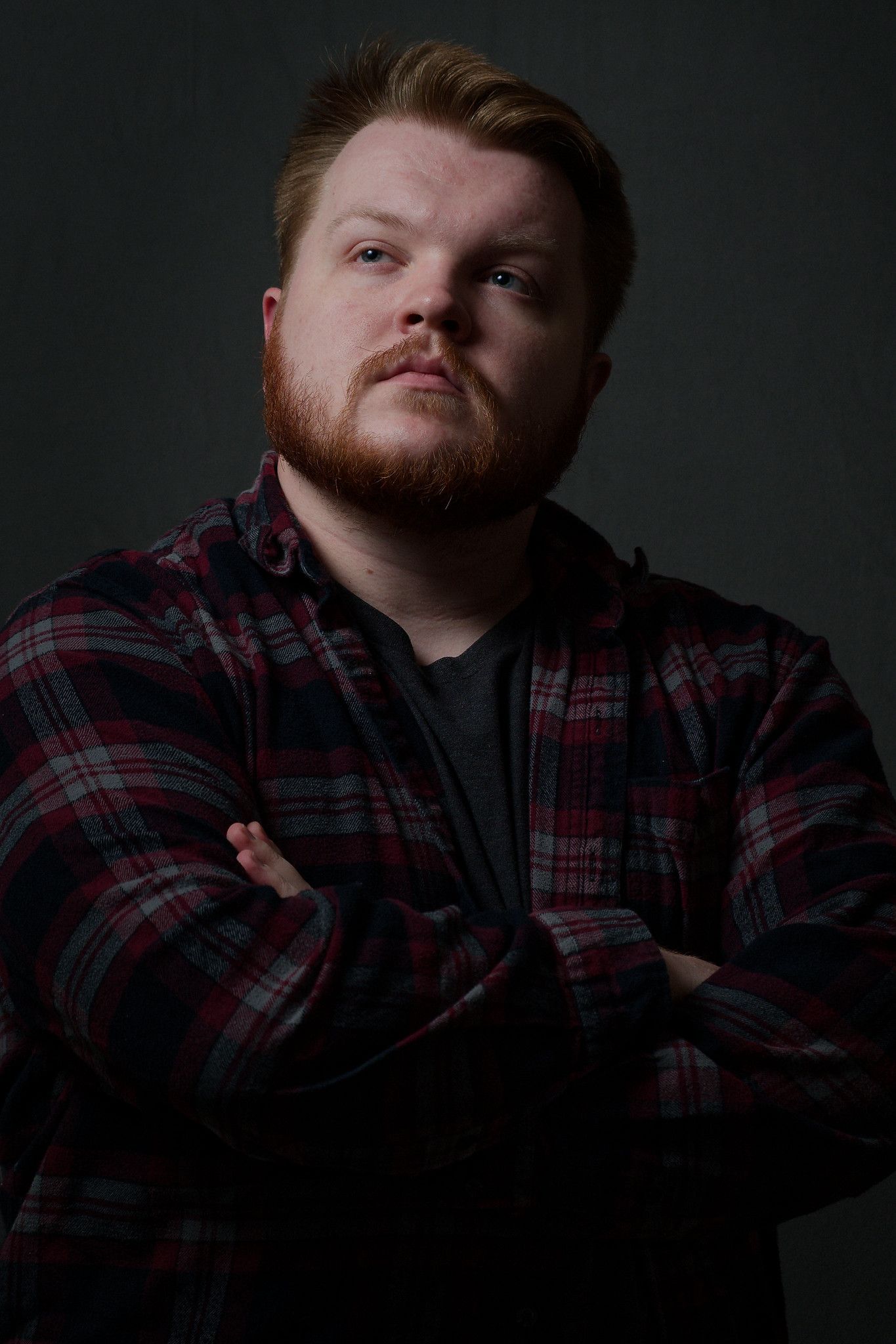Dramatic moody portrait of a man wearing a plaid flannel shirt and dark t-shirt. His arms are crossed.