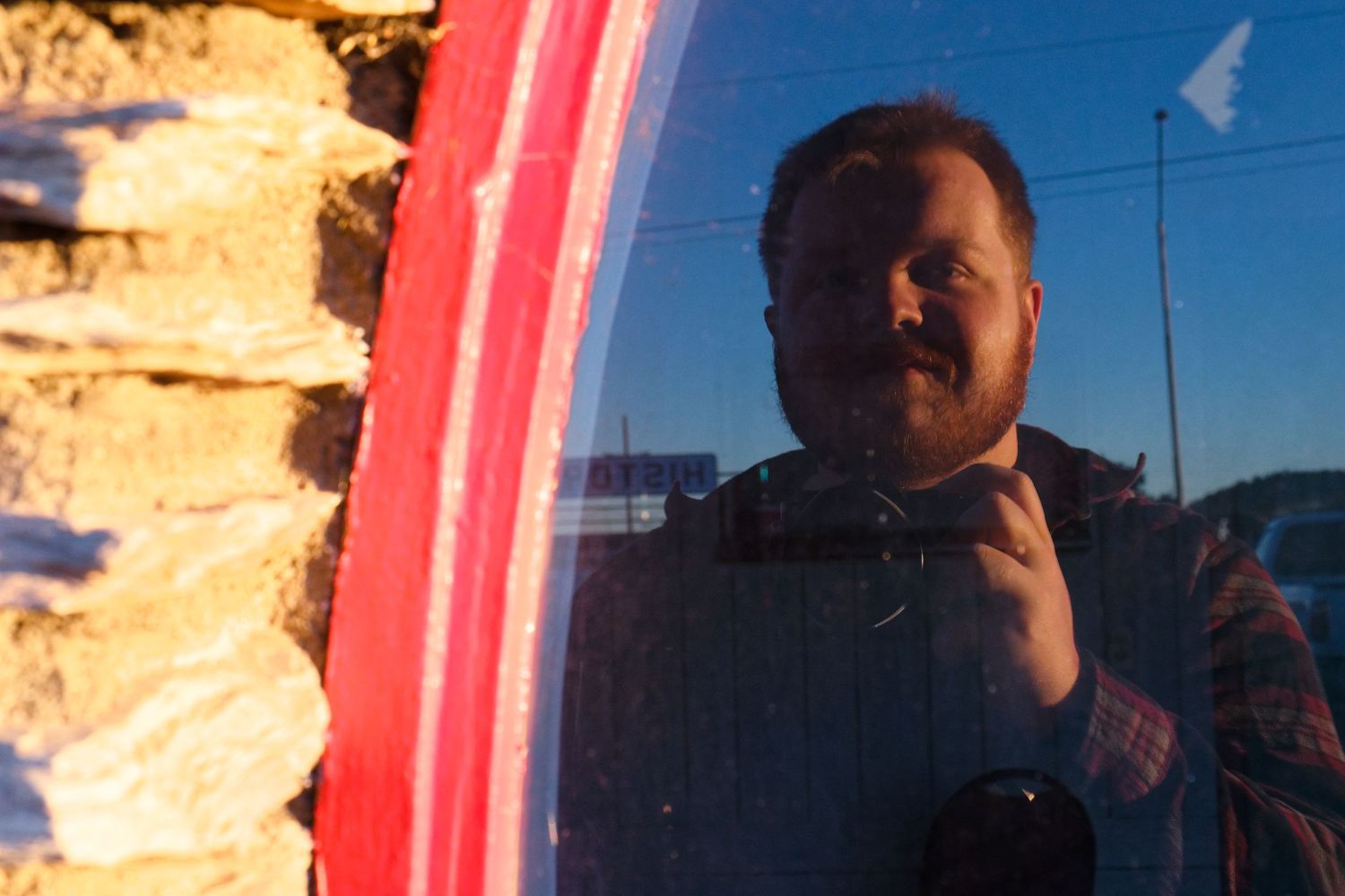 Reflected self portrait in a bright red surface with sunlit stone wall in background.
